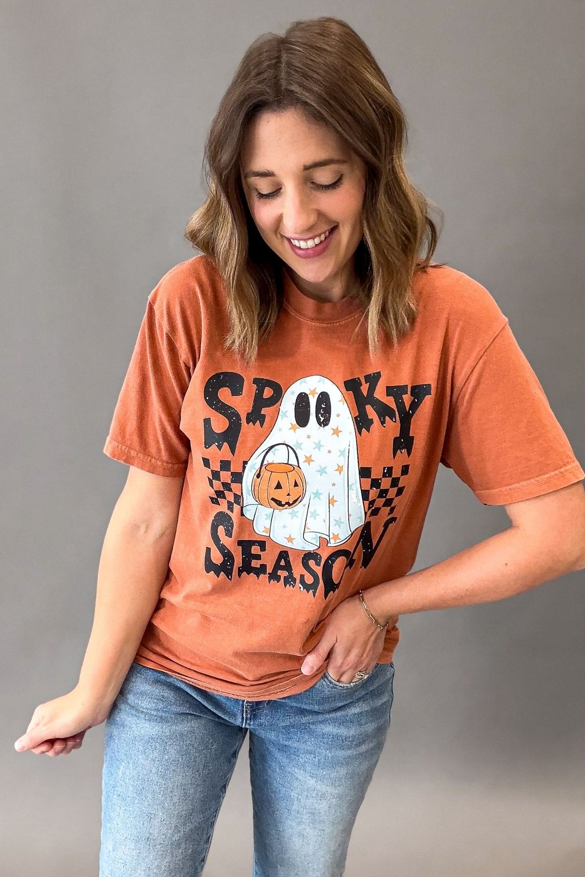 A woman wearing a burnt orange graphic t-shirt with a 'Spooky Season' Halloween design, paired with blue jeans, posing against a neutral gray studio background.