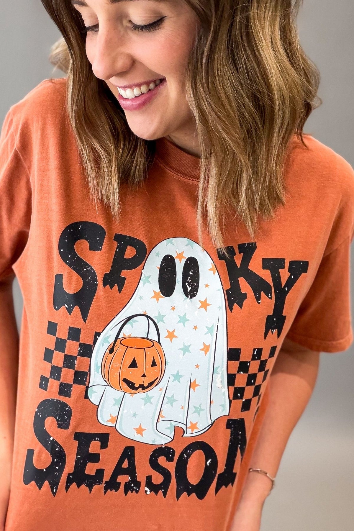 A woman wearing a burnt orange graphic t-shirt with a 'Spooky Season' Halloween design featuring a ghost, pumpkin, and stars.