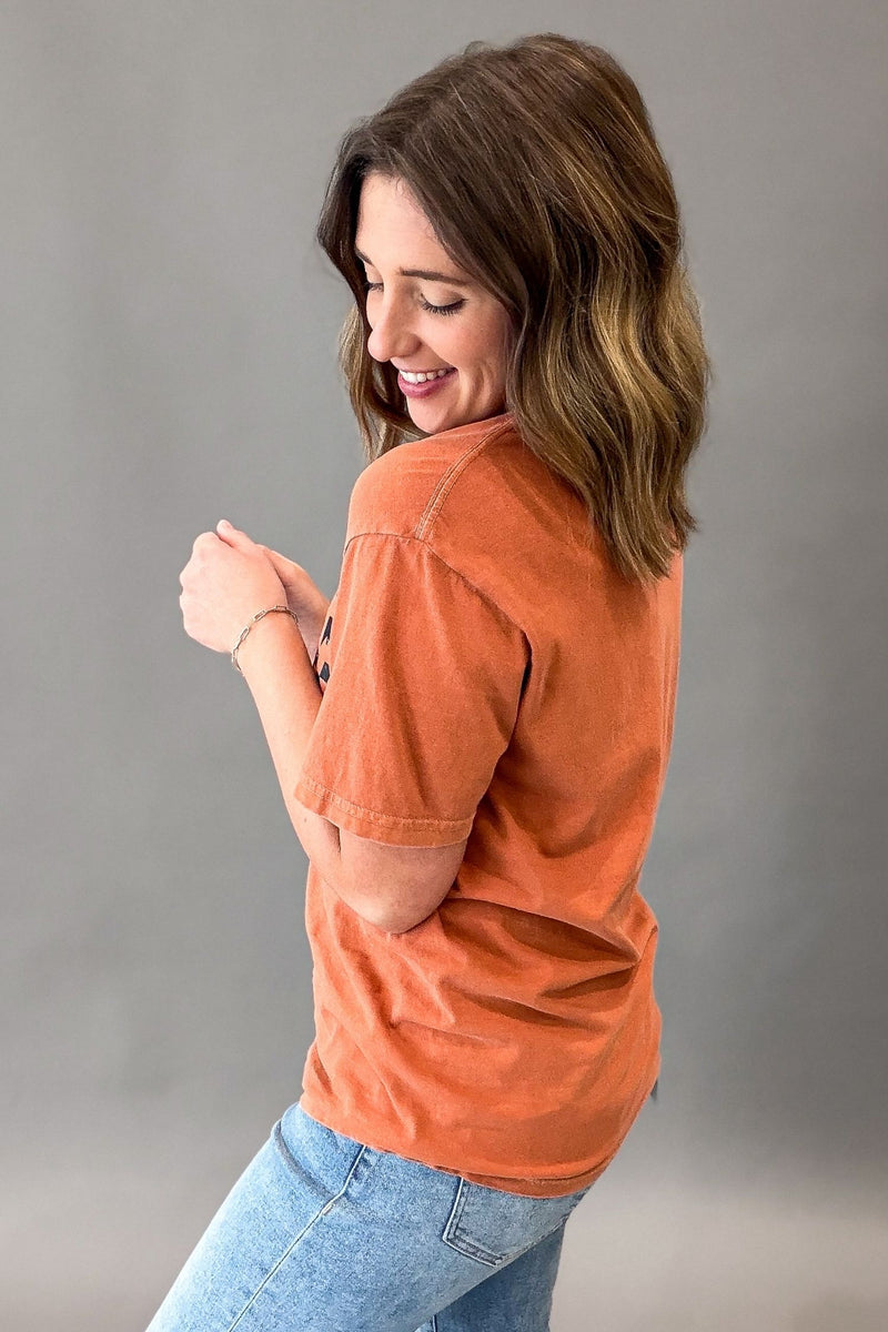 A woman wearing a washed orange short-sleeve t-shirt and light blue jeans, shown from a side-back angle against a neutral gray studio background.