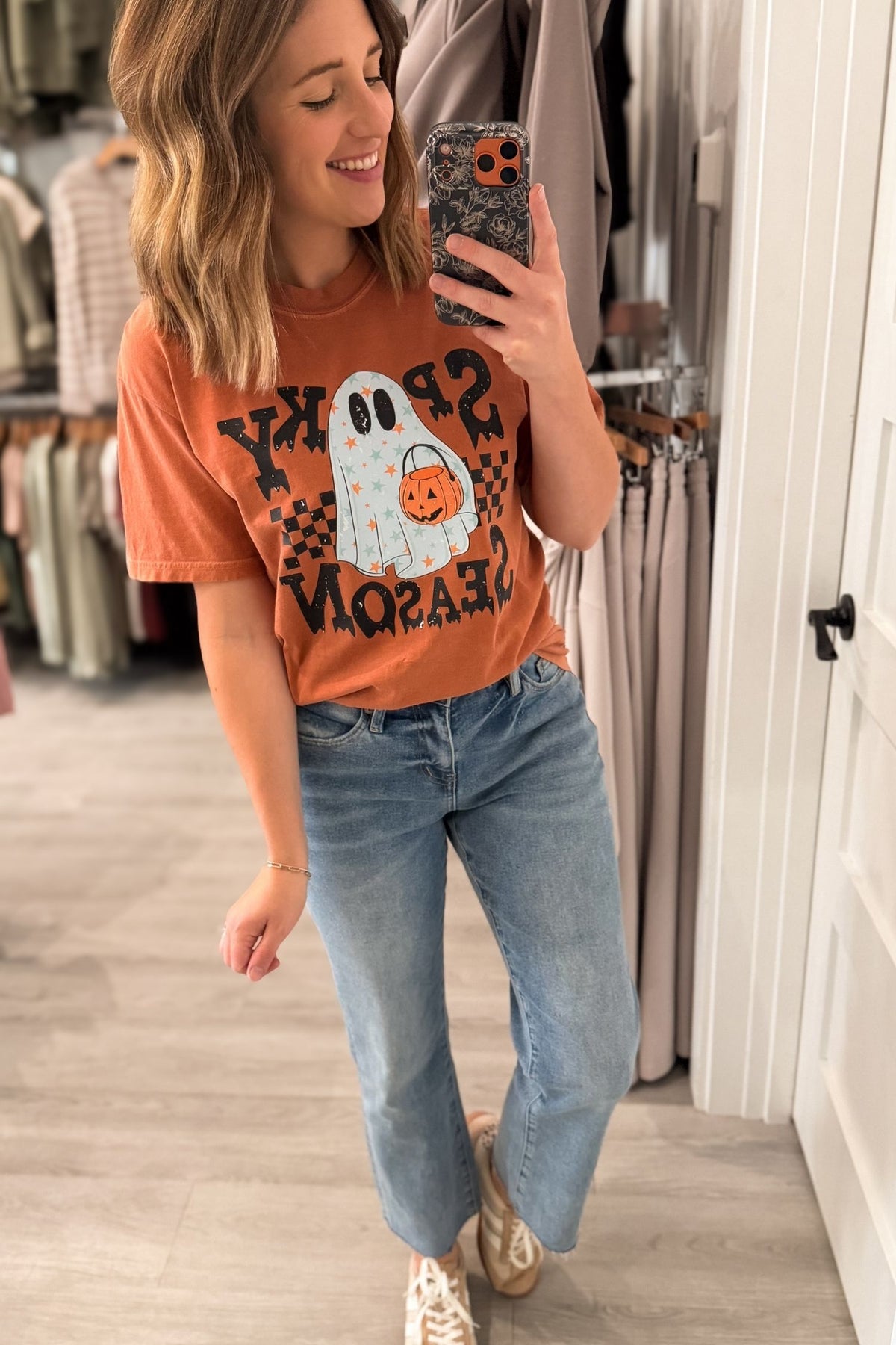 A woman takes a mirror selfie in a clothing store wearing a rust-orange graphic t-shirt with a Halloween ghost design, blue jeans, and beige sneakers.