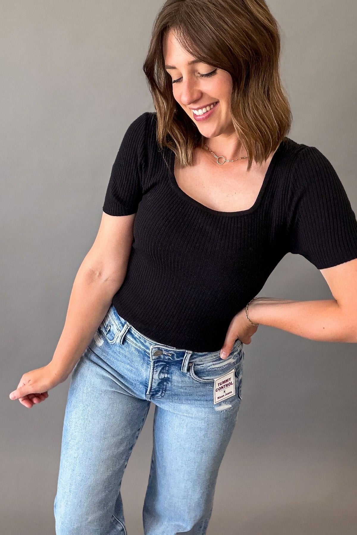 Woman wearing a black fitted ribbed square neck tank with blue jeans on a grey background