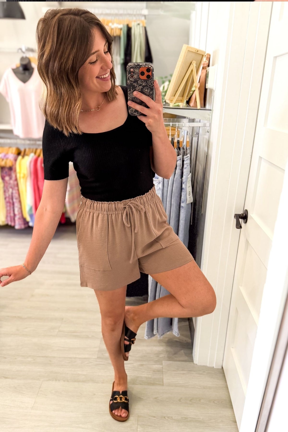 woman taking a mirror selfie in a clothing boutique wearing a black fitted ribbed square neck tank with blue jeans on a grey background