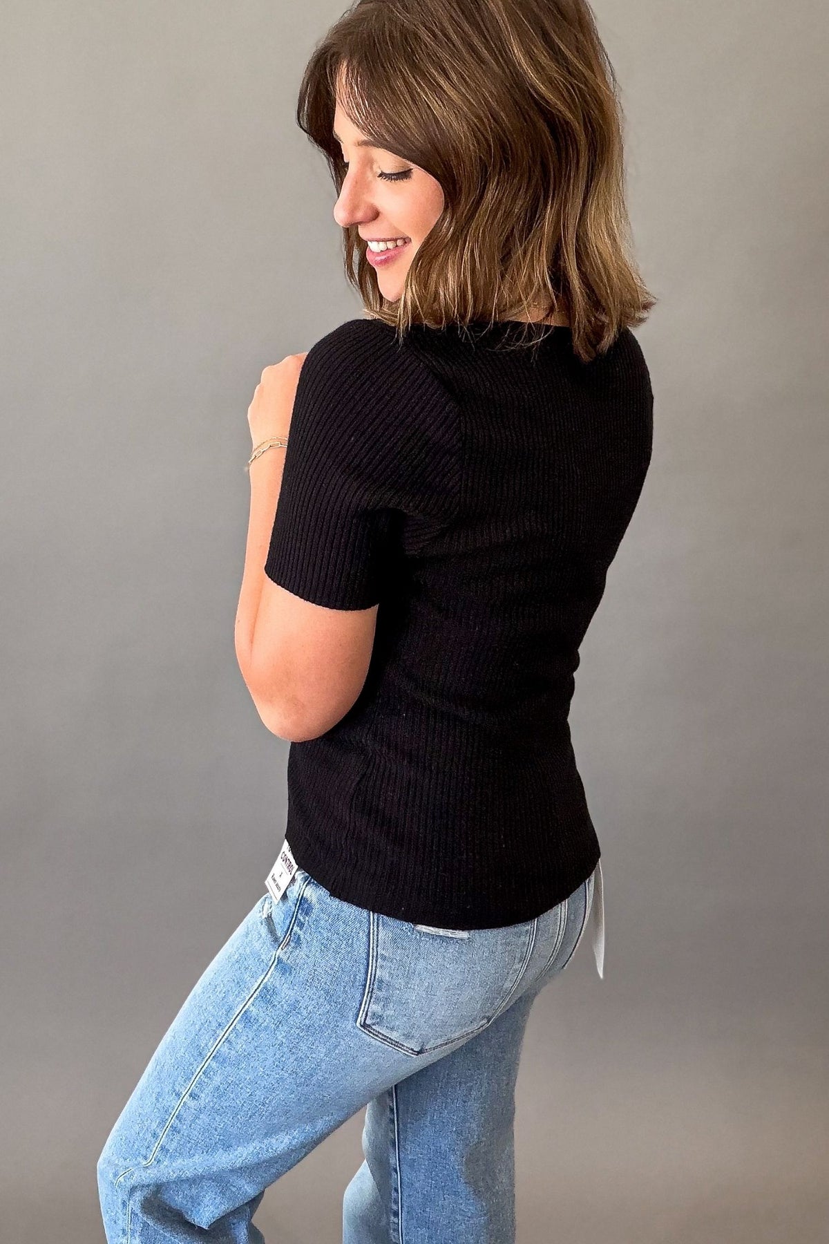 Woman wearing a black fitted ribbed square neck tank with blue jeans on a grey background showing the back and side details