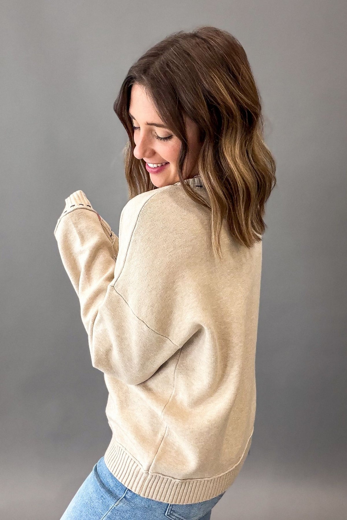 A woman with shoulder-length brown hair is shown from the back wearing a light beige knit sweater and blue jeans against a solid gray studio background.