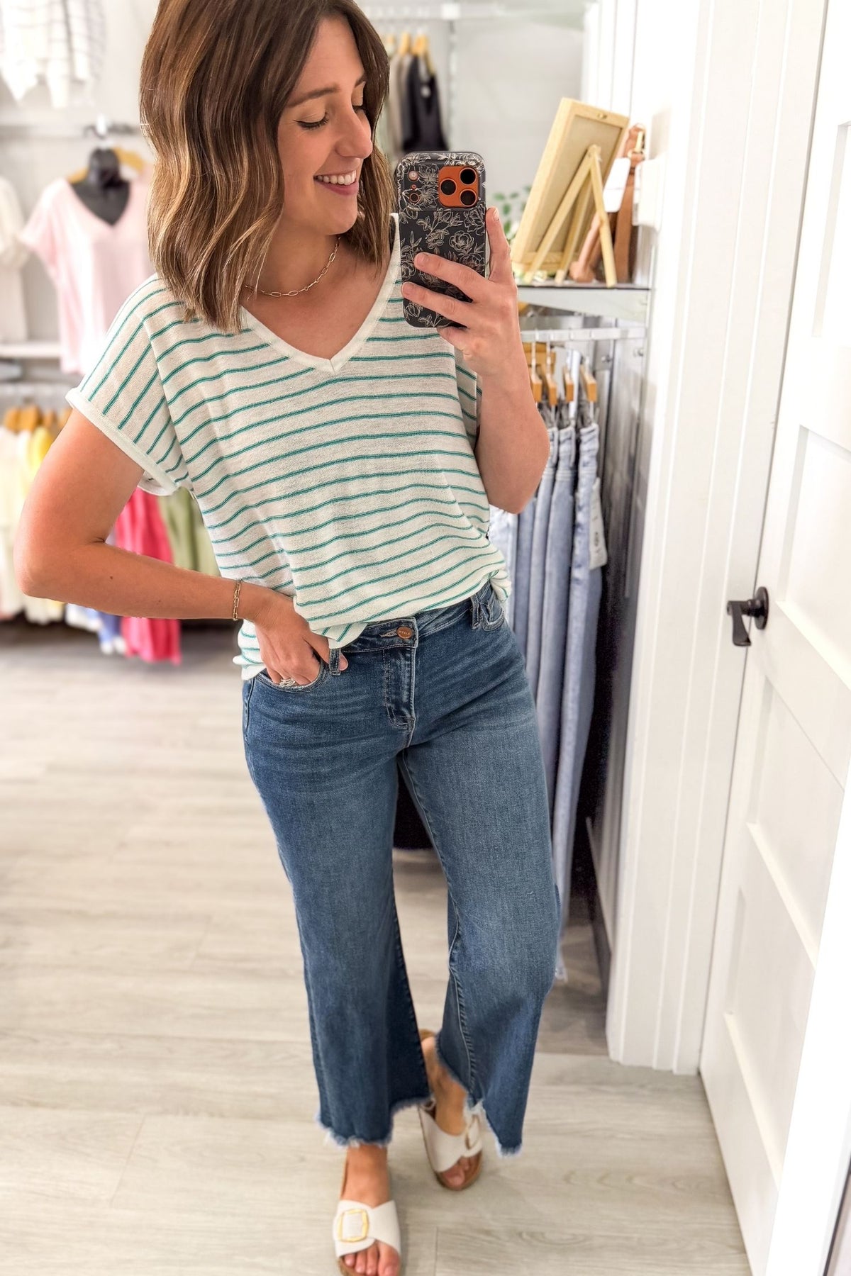Woman taking a mirror selfie wearing a teal striped shirt and jeans in a clothing store.