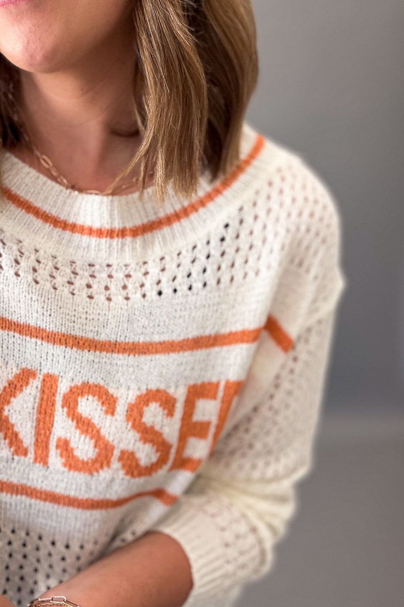 Person wearing a ivory sweater with orange text and stripes.