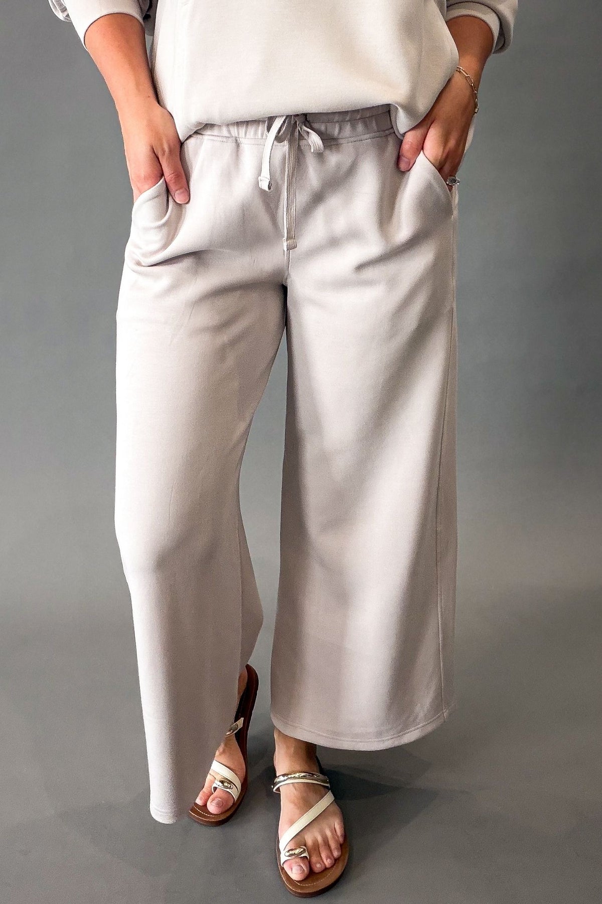 A person wearing mocha cream wide-leg drawstring pants with side pockets, paired with a cream sweatshirt and beige sandals, standing against a neutral gray studio background.