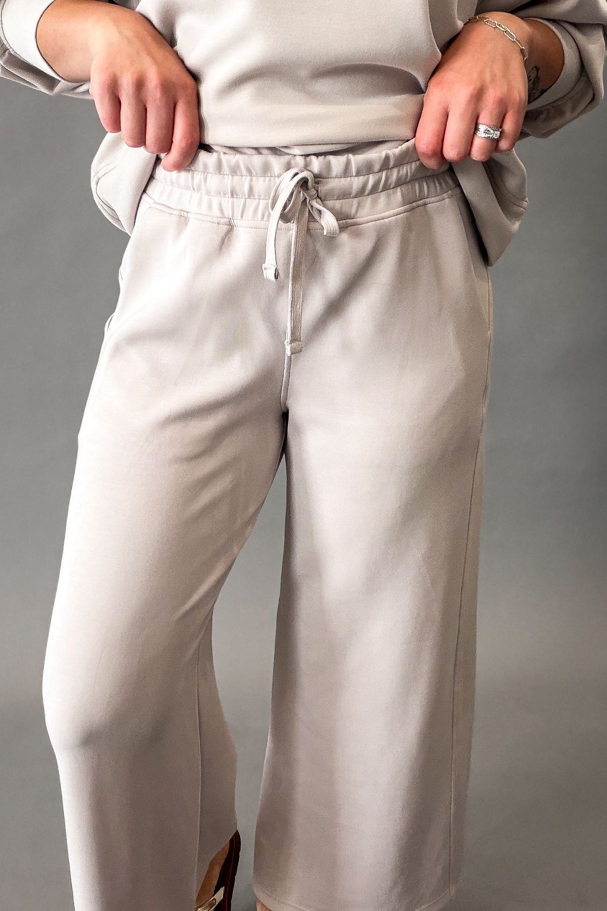 A person wearing  mocha cream wide-leg sweatpants with an elastic drawstring waist, paired with a matching beige top, shown against a plain gray studio background.