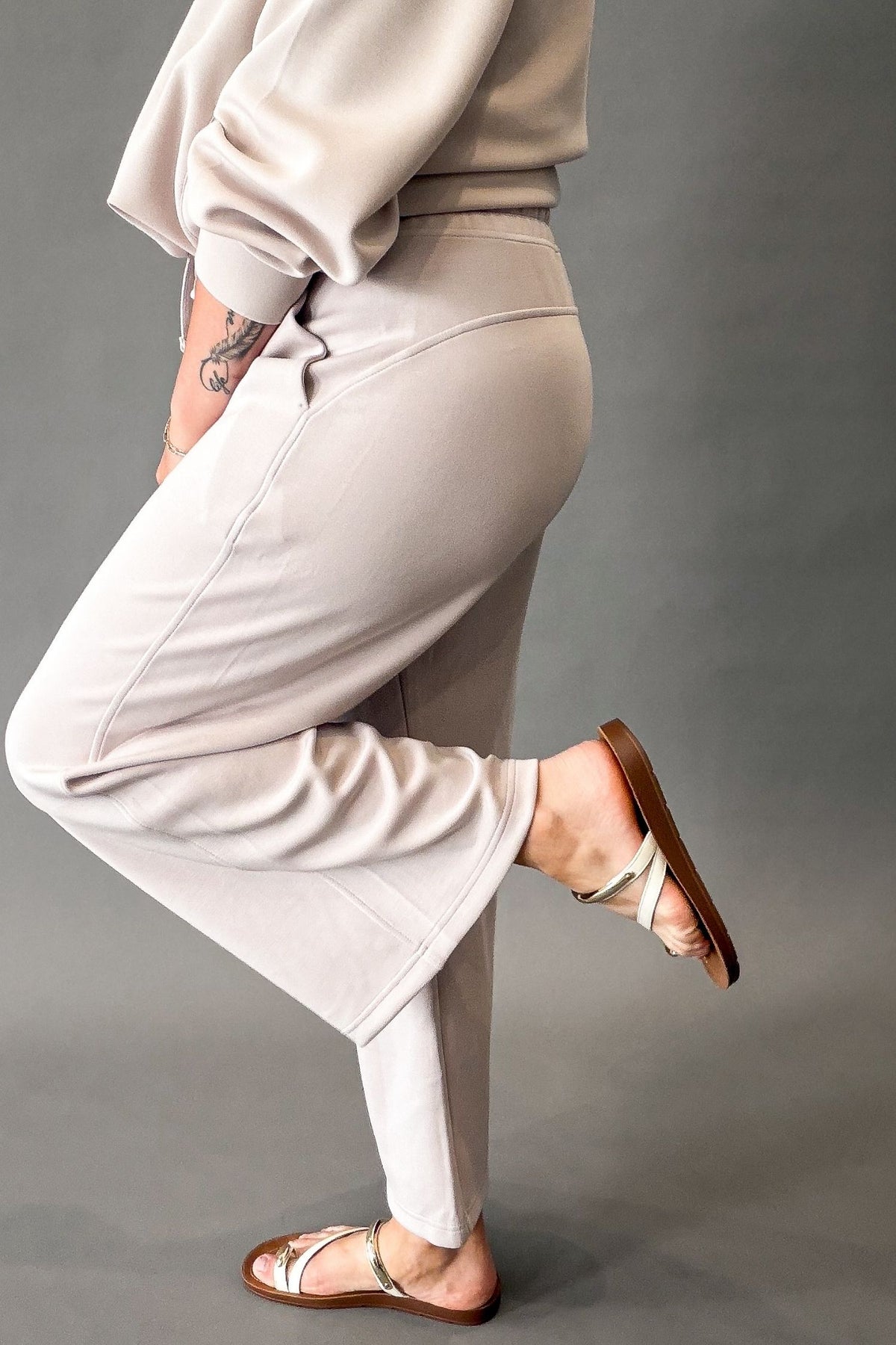 A person wearing a mocha cream lounge pants and a matching sweatshirt, shown from the side against a plain gray studio background.