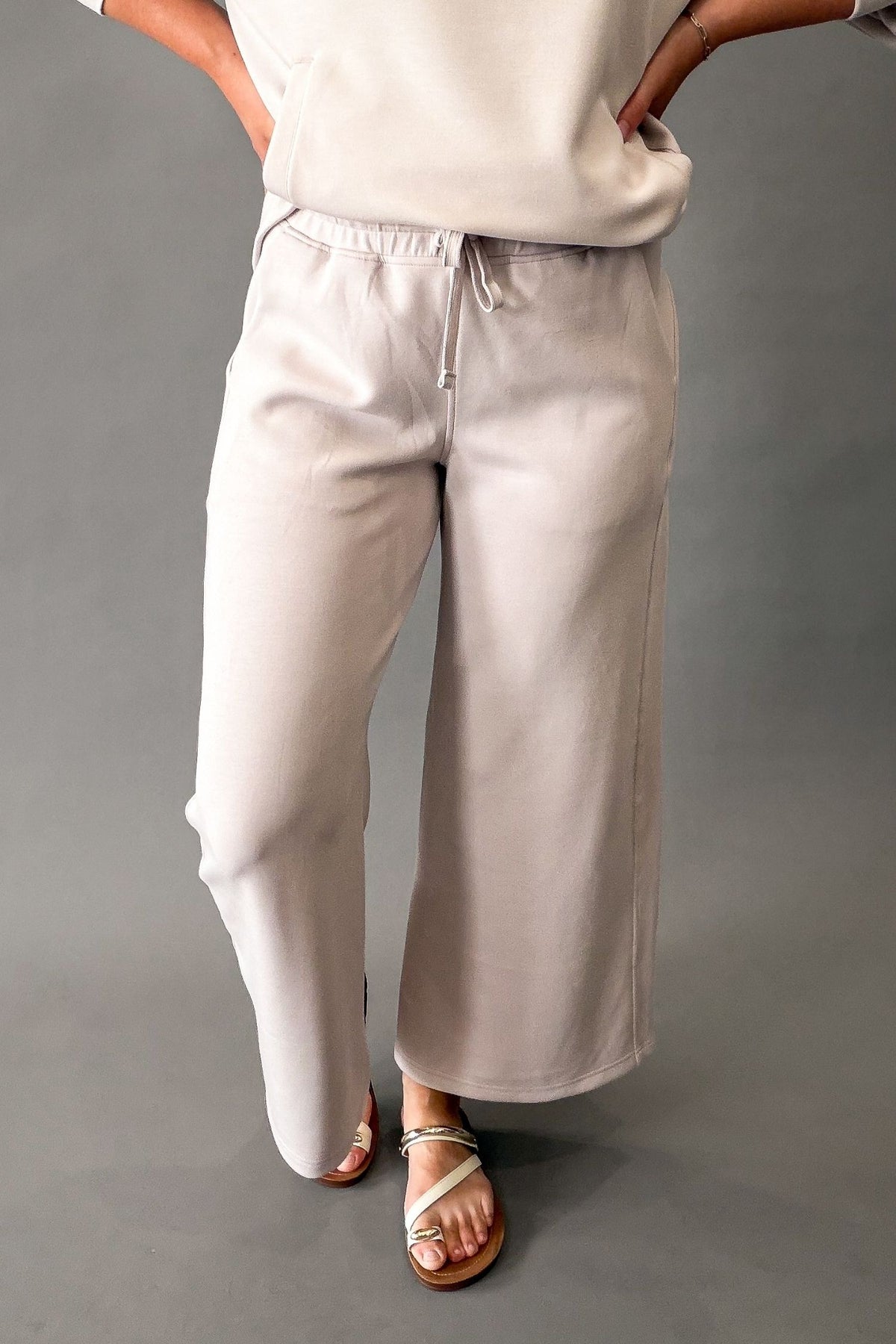 A person wearing a mocha cream wide-leg drawstring pants with a matching top and light colored sandals, standing against a plain gray studio background.