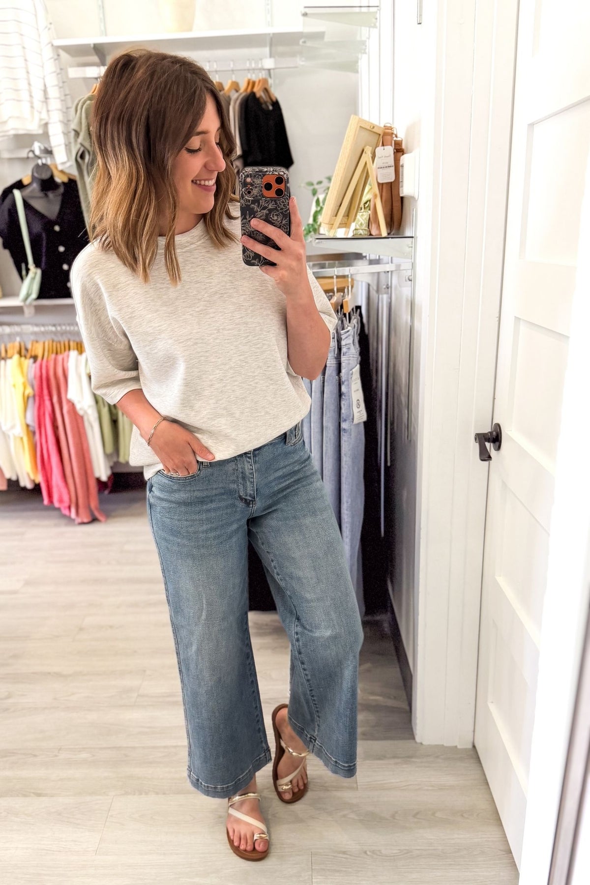 Woman taking a mirror selfie wearing a light heather grey short sleeve and blue jeans in a clothing store.