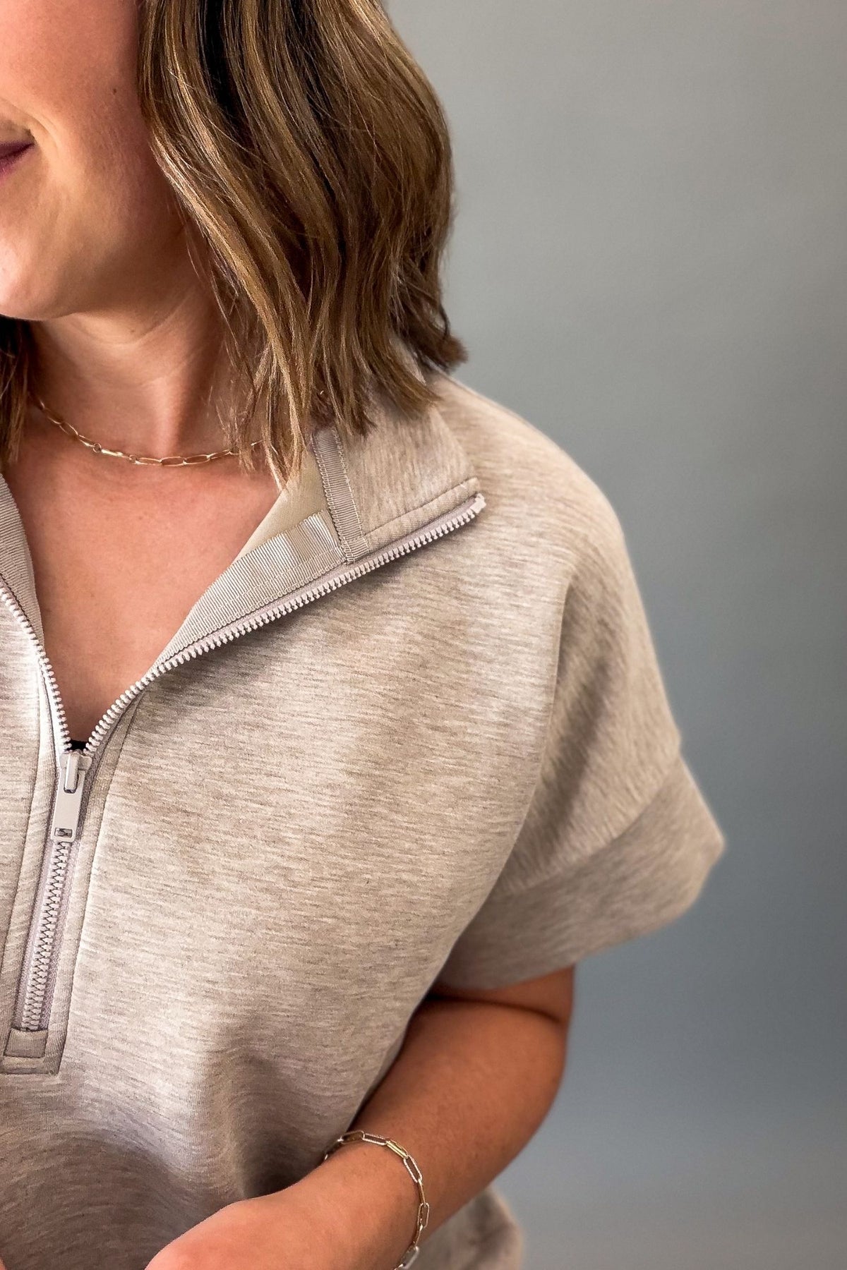 A close-up of a woman wearing a light heather taupe short-sleeve quarter-zip pullover, showing the collar, zipper, and part of her arm and hair against a plain gray background.