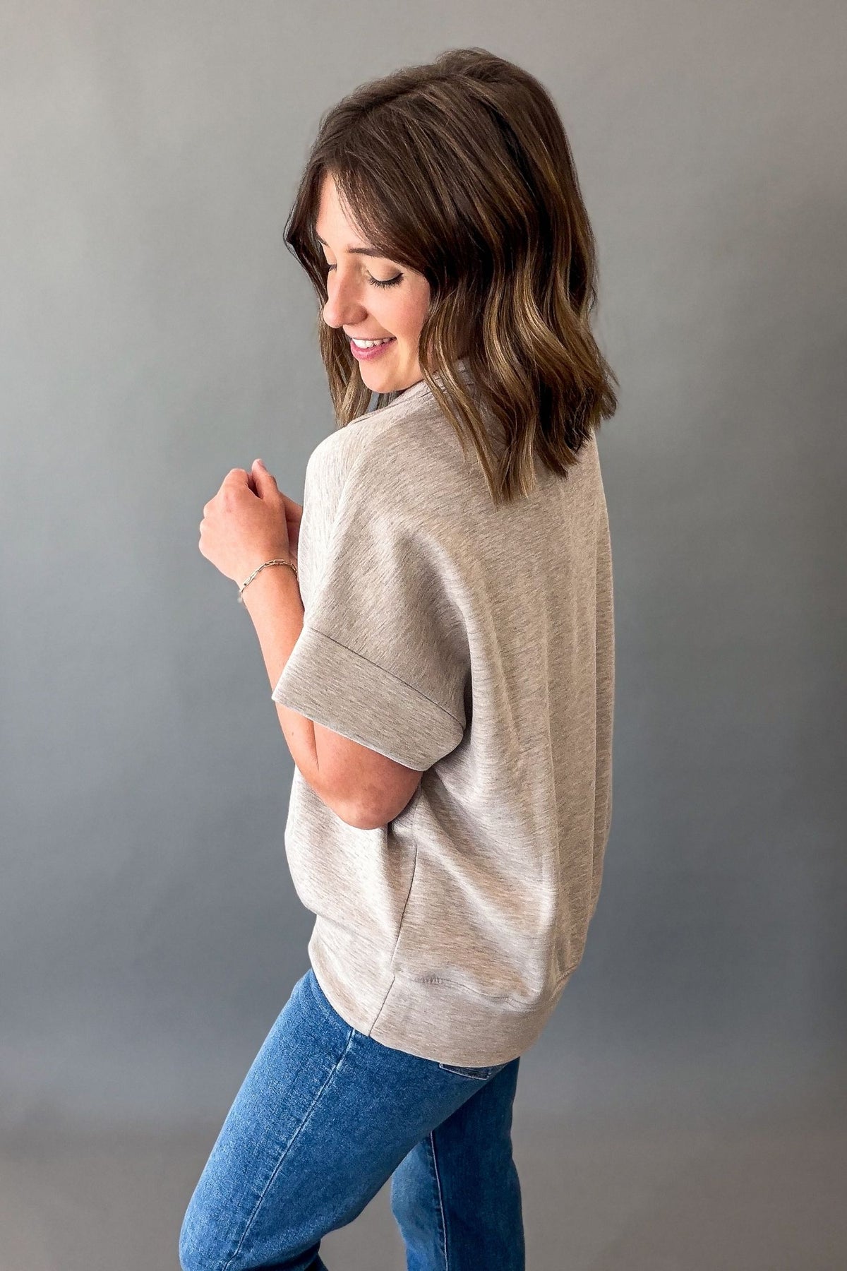 A woman wearing a light taupe heathered short-sleeve knit top and blue jeans, photographed from the side against a plain gray studio background.