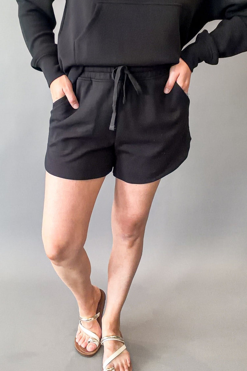 A person wearing black drawstring shorts with side pockets, paired with a black hoodie and beige flat sandals, standing against a plain gray studio background.