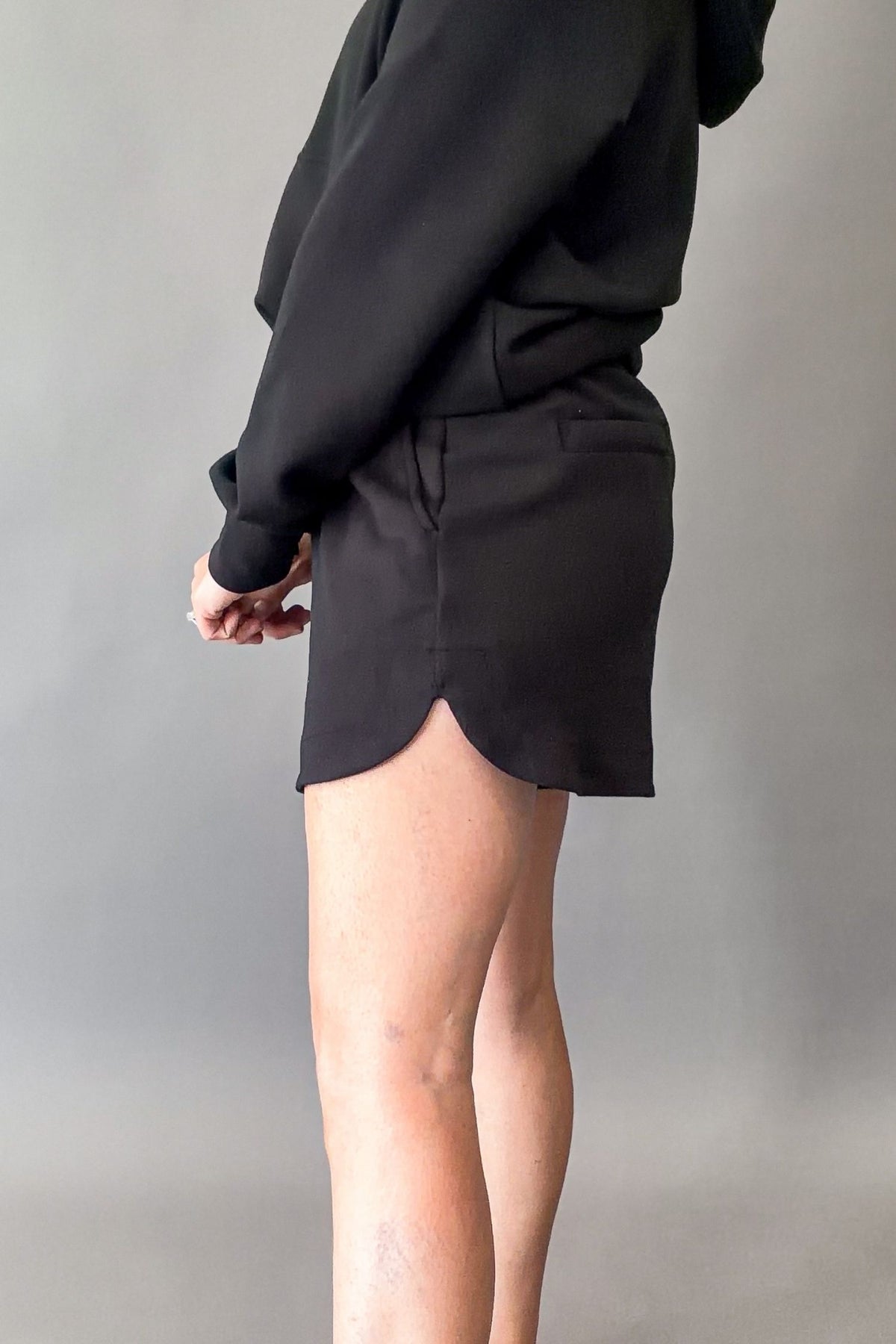 A person wearing a black oversized hoodie and a matching black shorts with a side slit hem, shown from the back against a plain gray studio background.