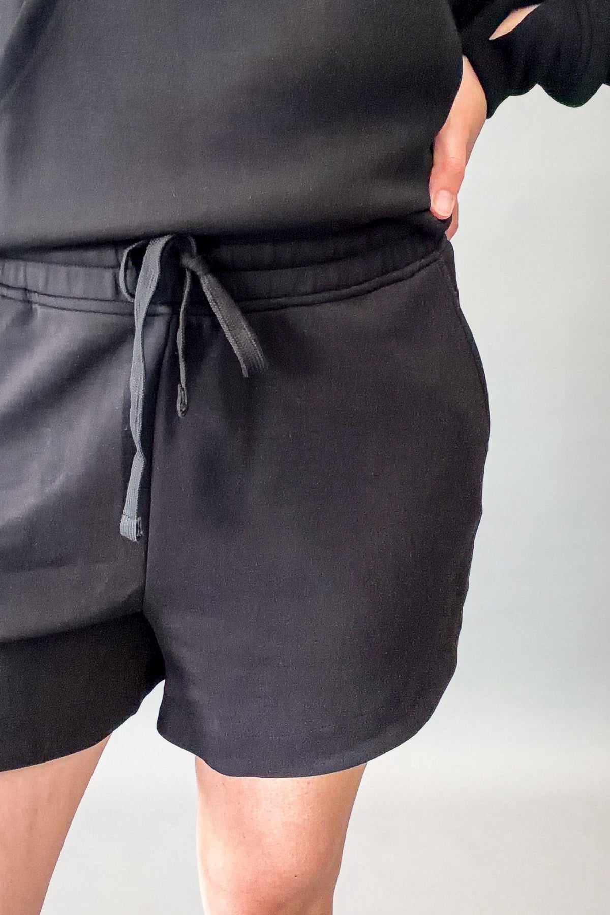 Close-up of a person wearing black drawstring shorts and a black sweatshirt against a light gray studio background.