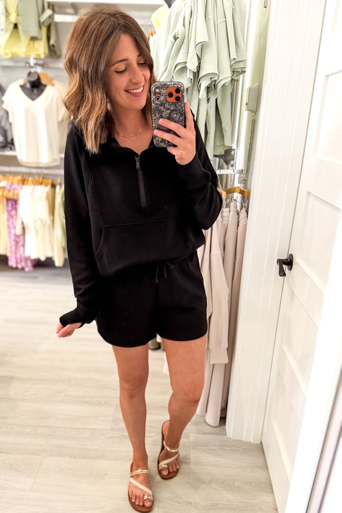 A woman takes a mirror selfie in a clothing boutique wearing a matching black lounge set consisting of a half-zip long-sleeve top and shorts, paired with beige strappy sandals.