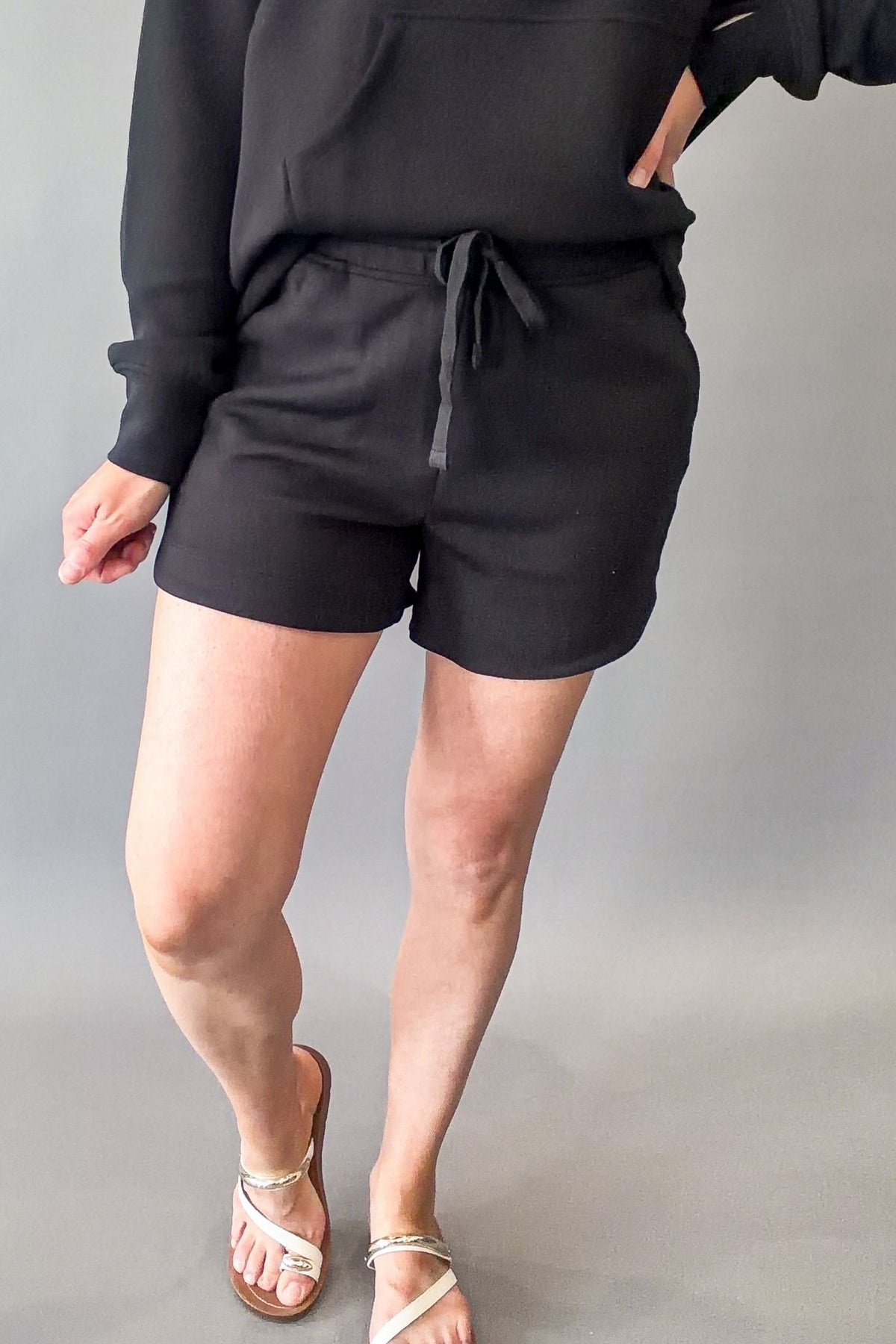 A person wearing black drawstring shorts with a black sweatshirt and beige strappy sandals, standing against a plain gray studio background.