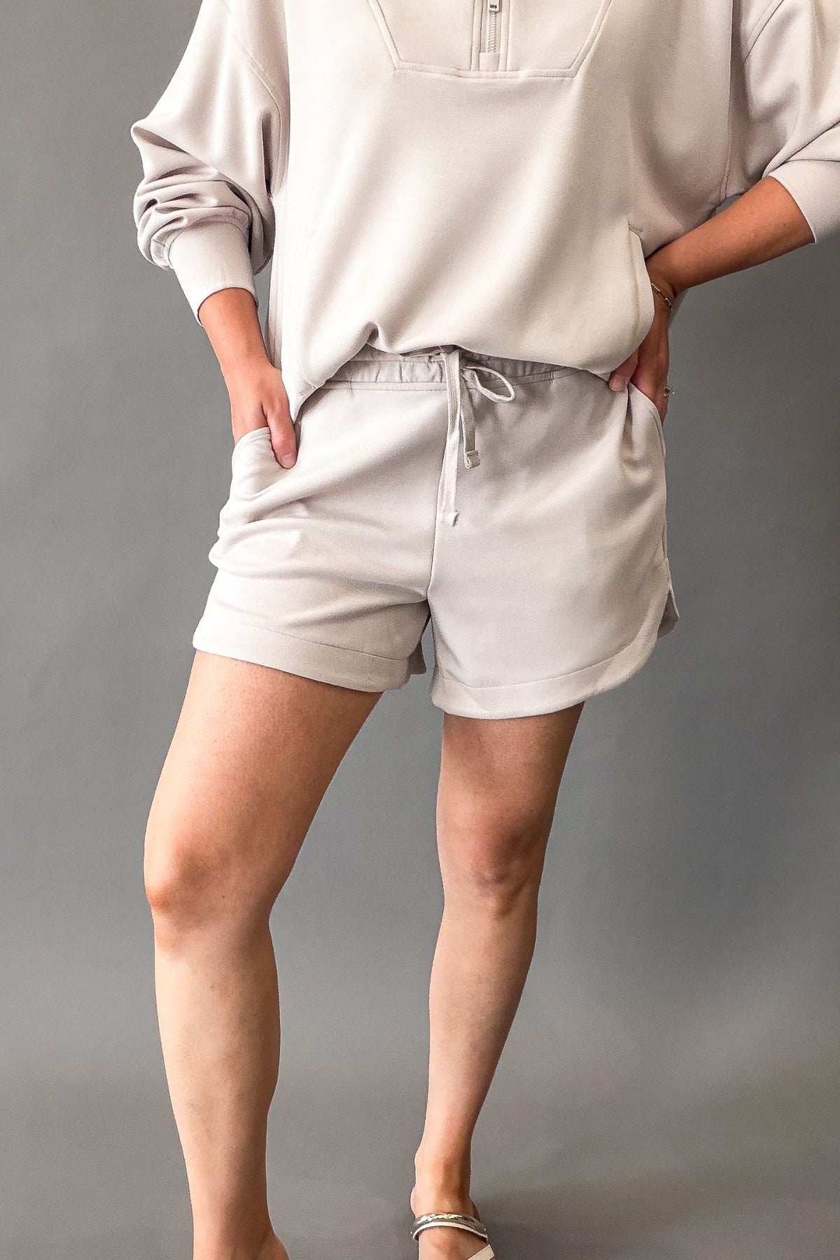 A model wearing light mocha cream drawstring shorts and a matching light beige long-sleeve top, standing against a neutral gray studio background.