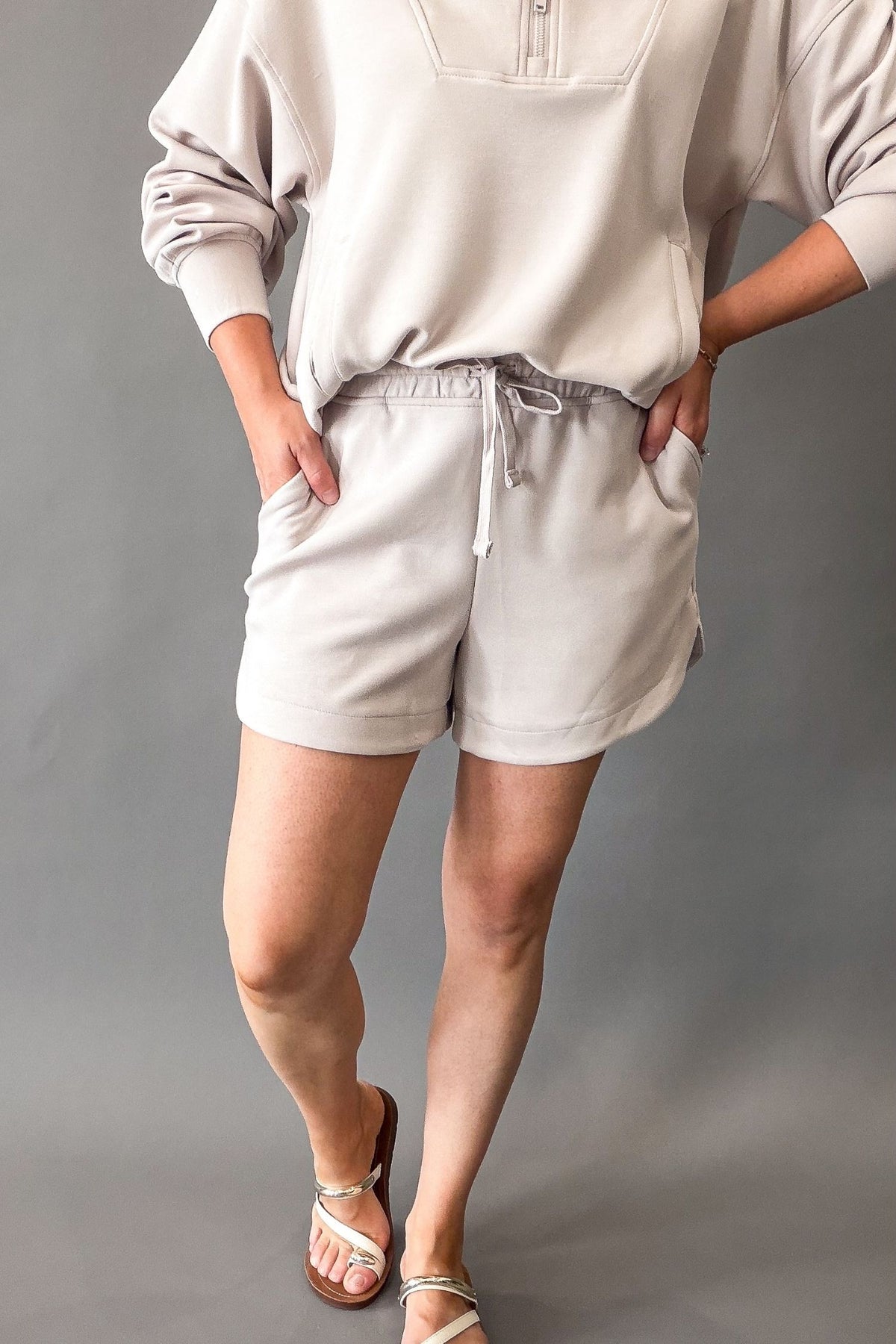 A model wearing light mocha cream lounge shorts with a drawstring waist and a matching beige sweatshirt, paired with off white strappy sandals, against a solid gray studio background.
