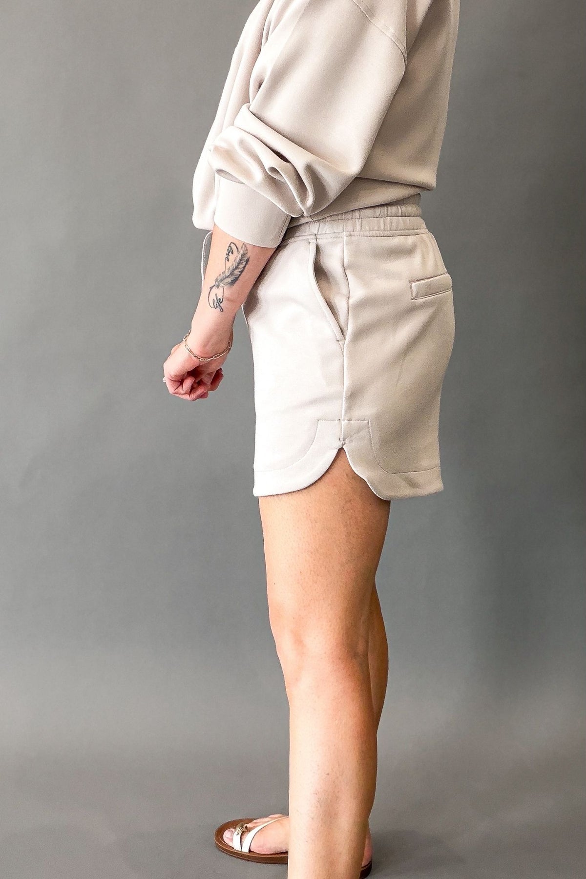 A person shown from the back wearing a light mocha cream sweatshirt and matching high-waisted shorts with side pockets, standing against a plain gray studio background.