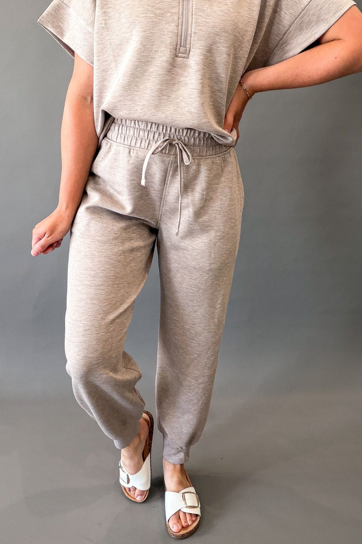 A person modeling a light heather taupe lounge set consisting of a short-sleeve half-zip top and matching drawstring sweatpants, paired with white slide sandals against a solid gray studio background.