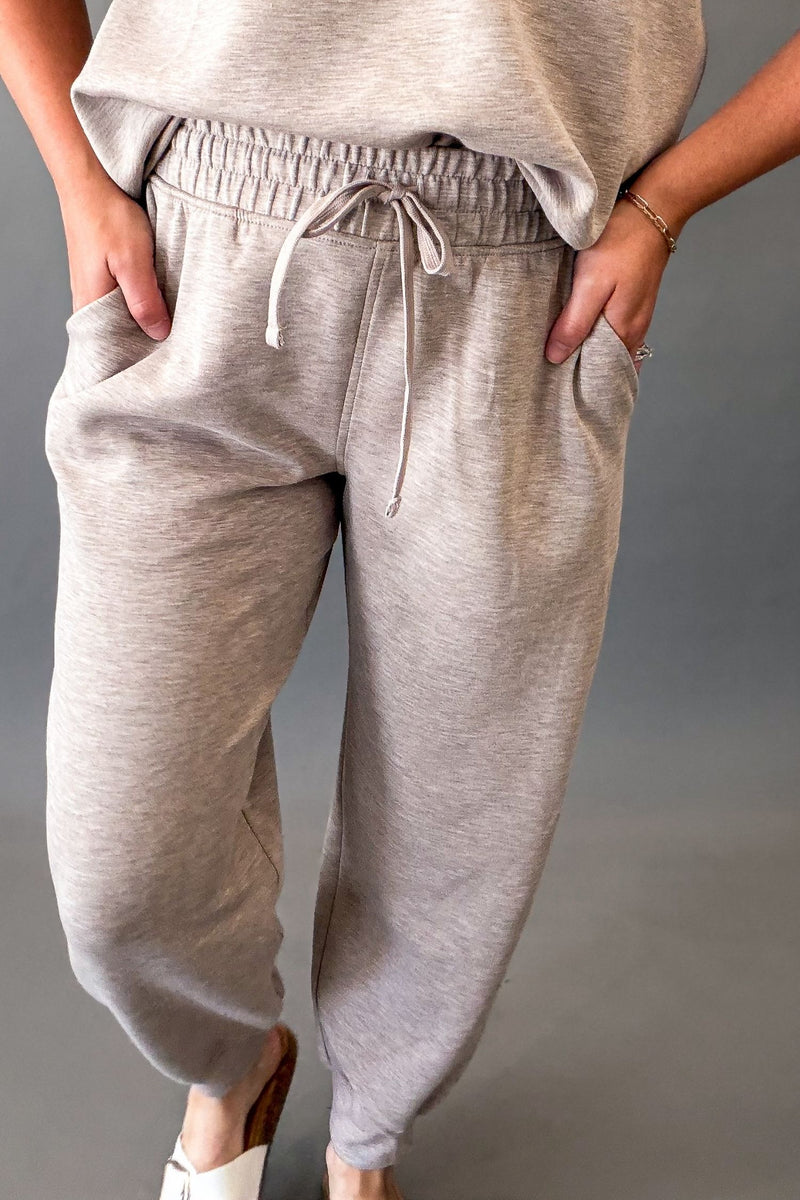 A person wearing light heather taupe jogger sweatpants with an elastic drawstring waist and side pockets, paired with a matching top and white slide sandals, against a plain gray studio background.