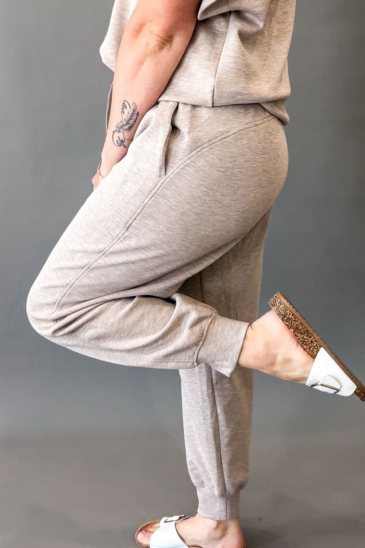 A person wearing heather taupe jogger sweatpants and a matching top, shown from the waist down against a plain gray studio background. The person is wearing white slide sandals with leopard-print soles.