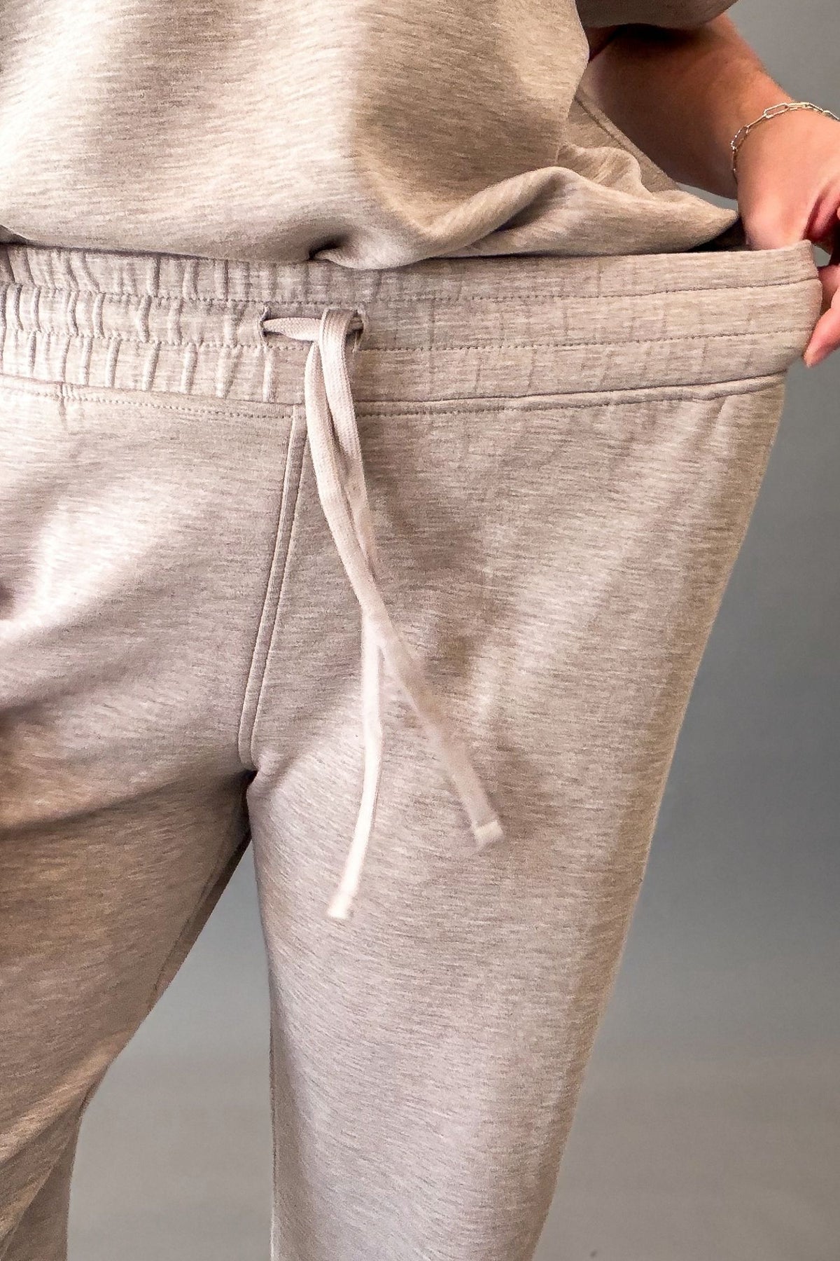 Close-up of a person wearing heather taupe jogger sweatpants with an elastic drawstring waist, holding the side of the pants to show the fit.