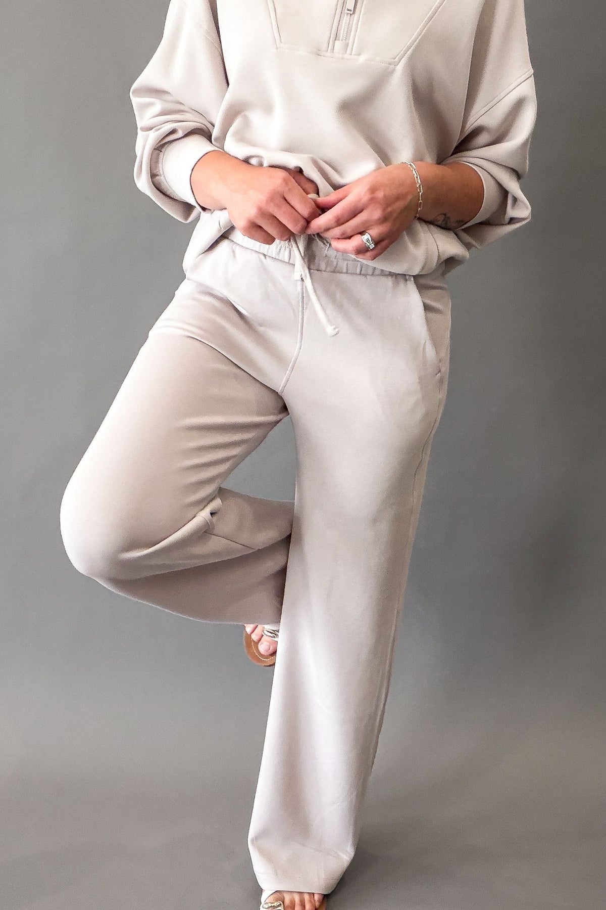 A person wearing soft mocha lounge pants and a matching sweatshirt, standing against a plain gray studio background.