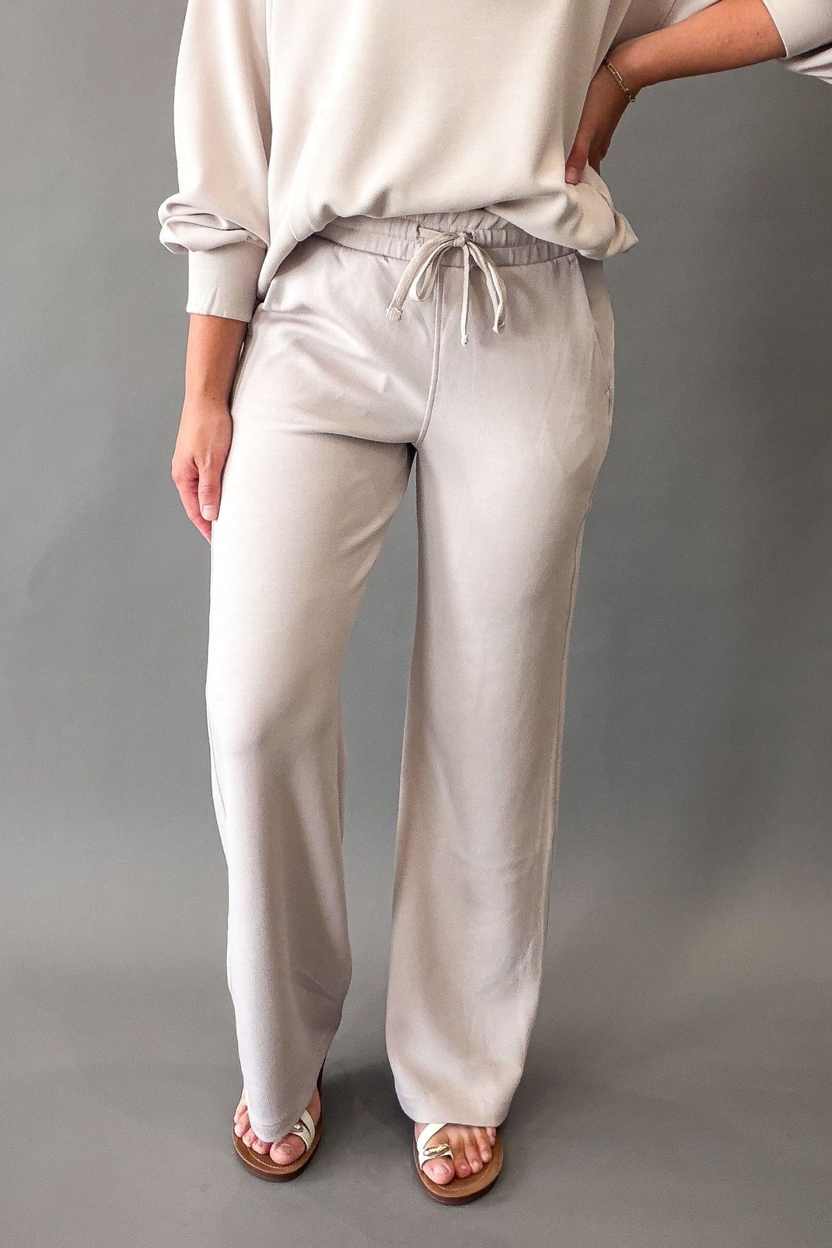 A person wearing soft mocha straight leg drawstring pants with a matching sweatshirt, shown from the waist down against a plain gray studio background.