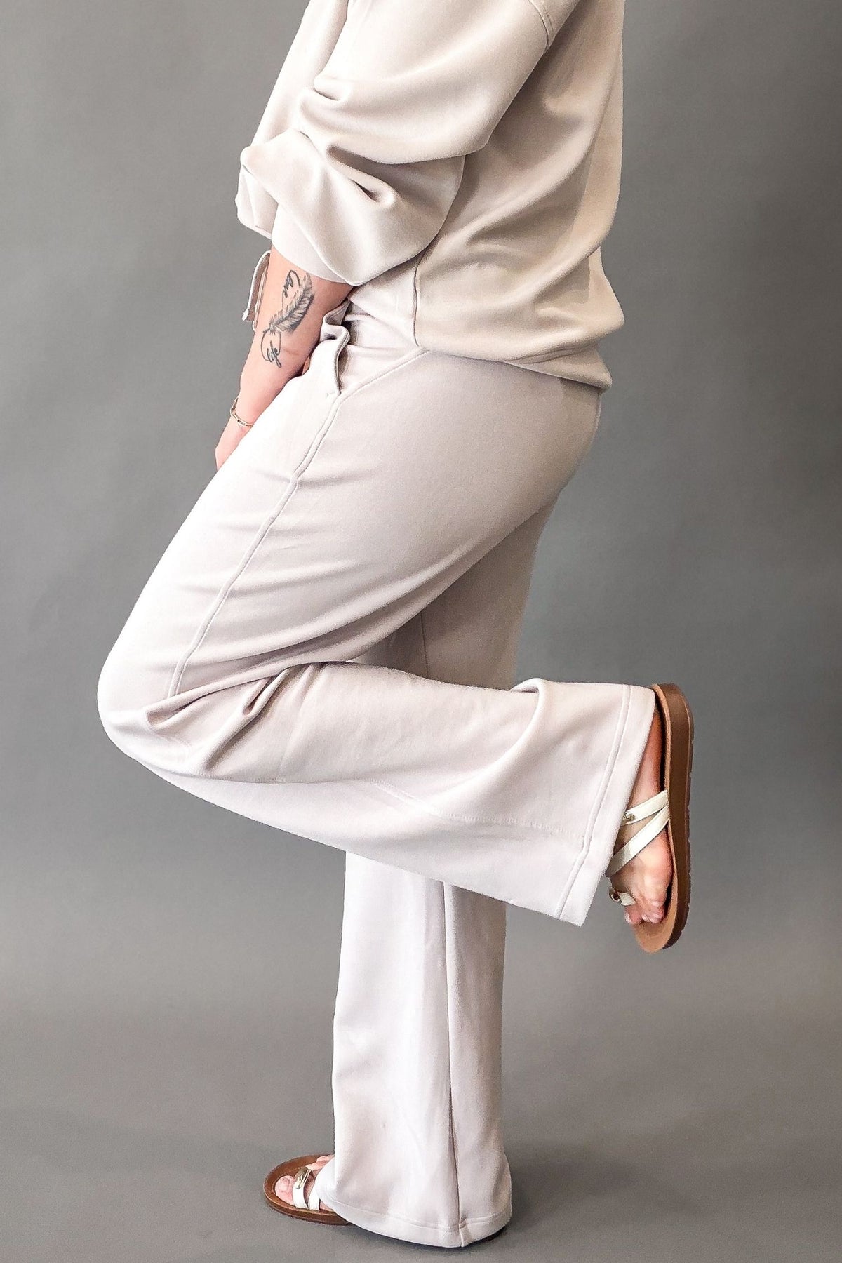 A person wearing soft mocha straight pants and a matching beige top, shown from the waist down in a studio setting against a neutral gray background.