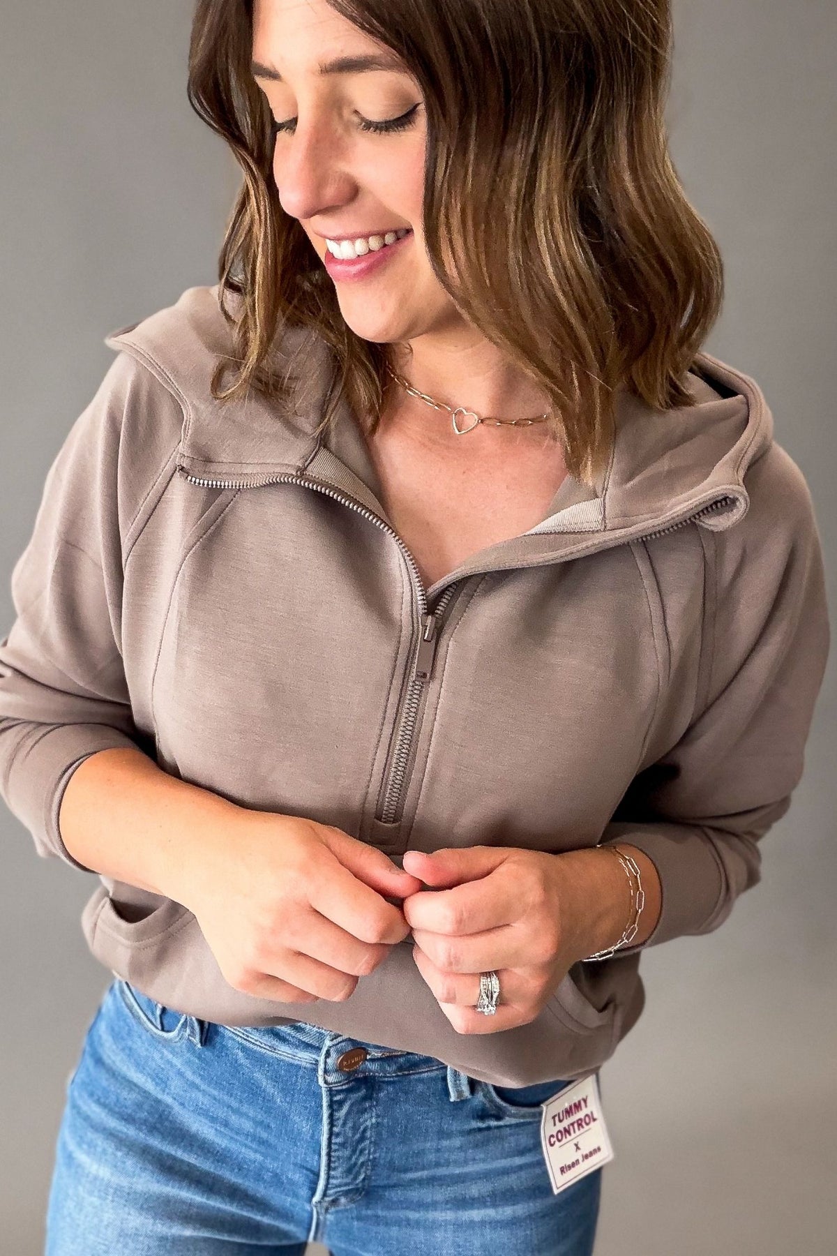 A woman wearing a mocha-brown half-zip hoodie with a hood and kangaroo pocket, paired with blue jeans, shown against a neutral gray background.