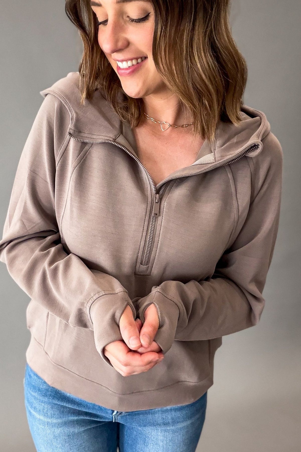A woman wearing a mocha brown half-zip hoodie with a split neckline and ribbed cuffs, paired with blue jeans, photographed against a neutral gray background.