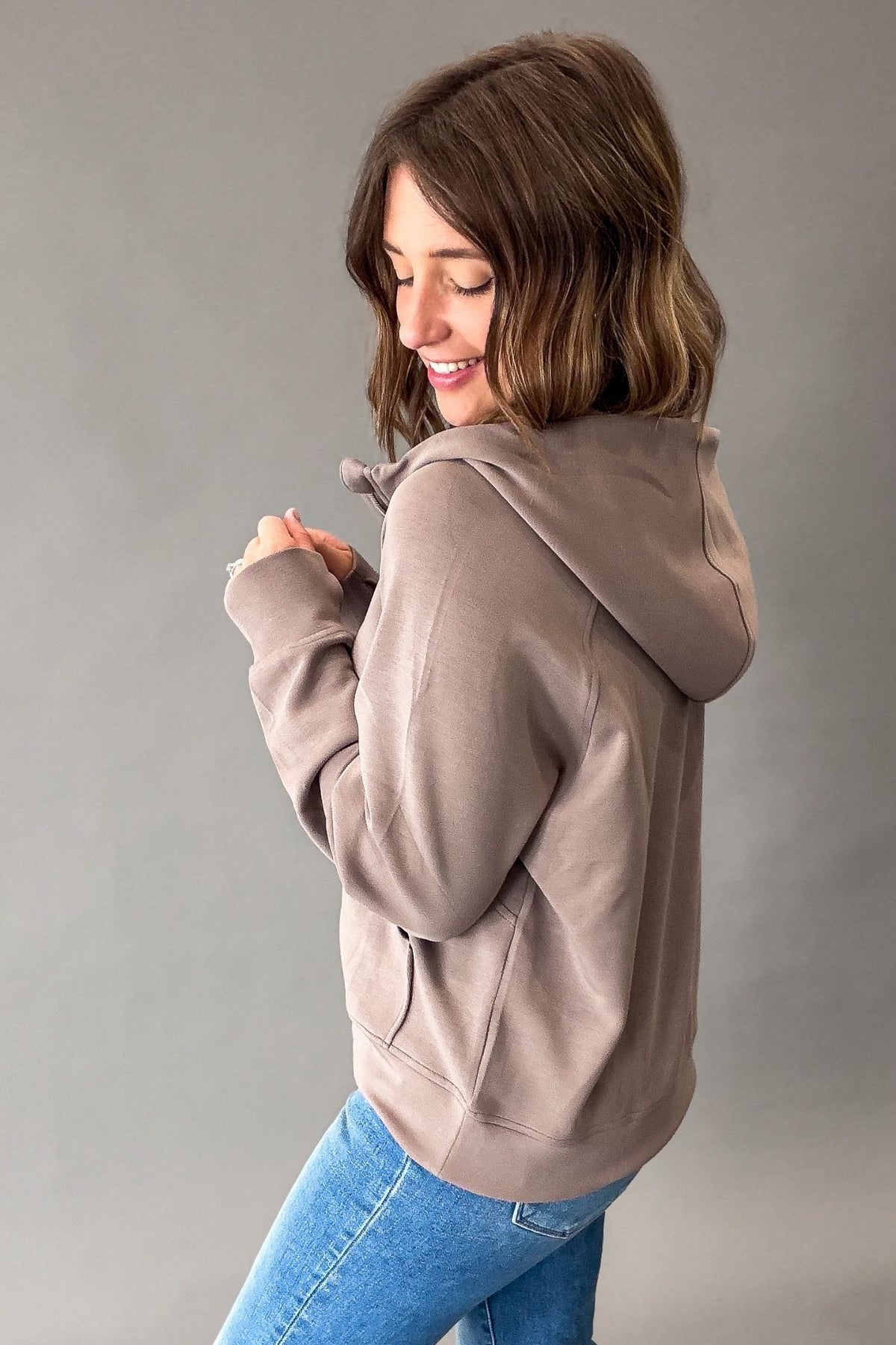 A woman with shoulder-length brown hair is shown from the back wearing a mocha-brown pullover hoodie and blue jeans against a solid gray studio background.