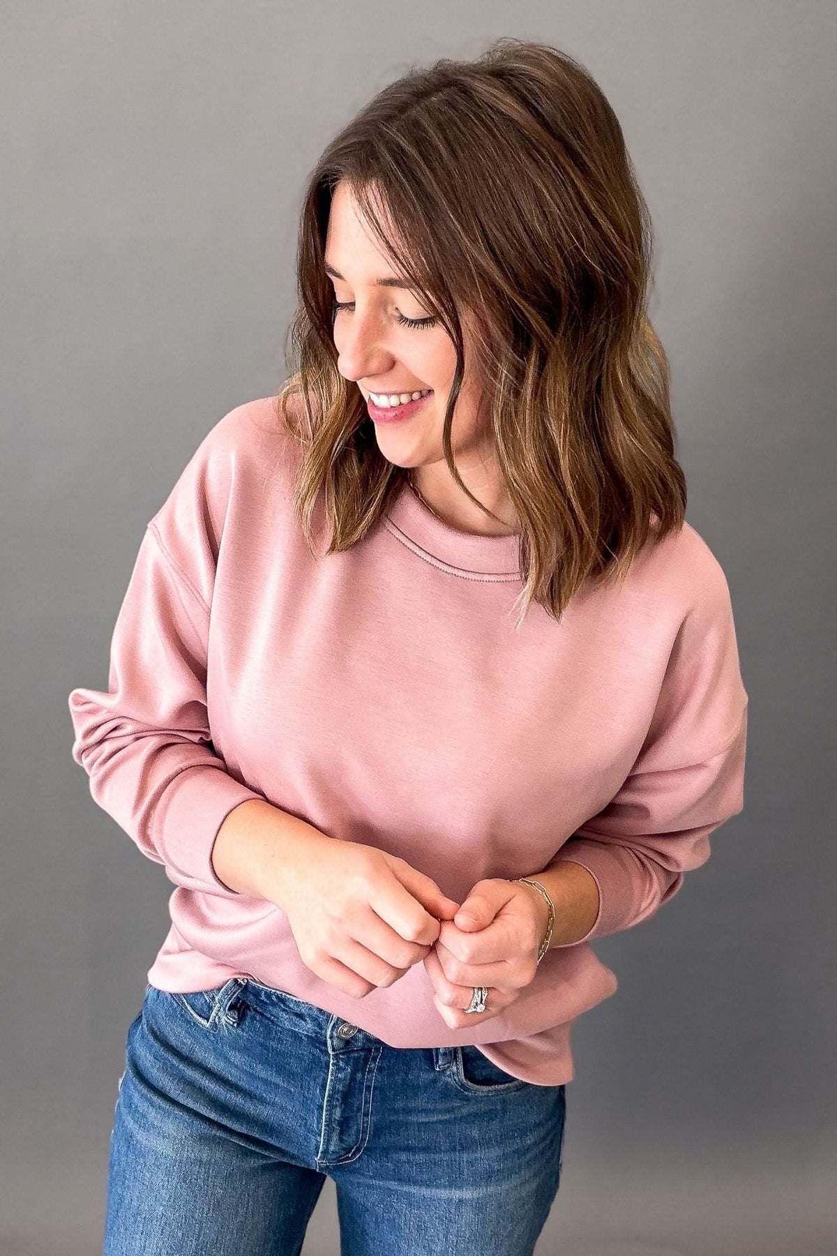 A woman with shoulder-length brown hair is wearing a dusty pink crewneck sweatshirt and blue jeans, standing against a solid gray studio background.