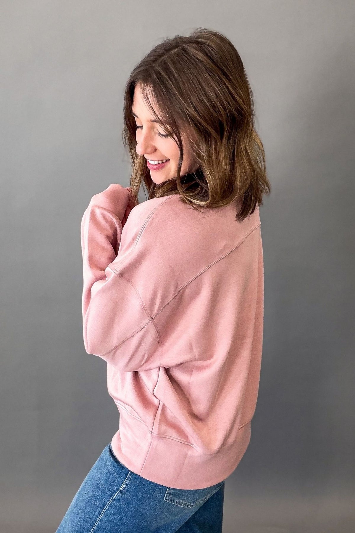 A woman with shoulder-length brown hair is shown from the back wearing a dusty pink crewneck sweatshirt and blue jeans, posing against a solid gray studio background.