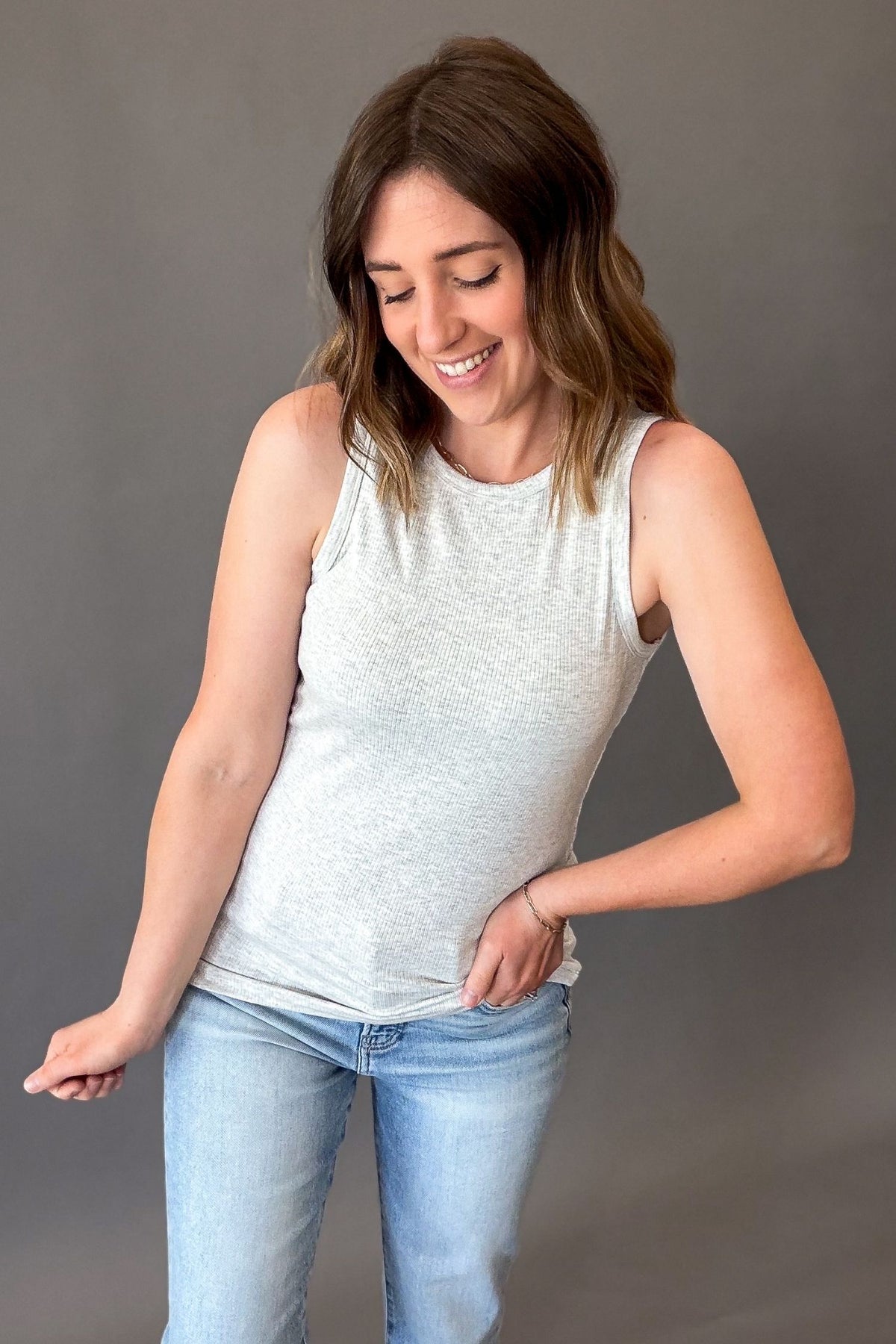 woman wearing a heather grey tank untucked to show length, has ribbed texture, with blue jeans on a grey background