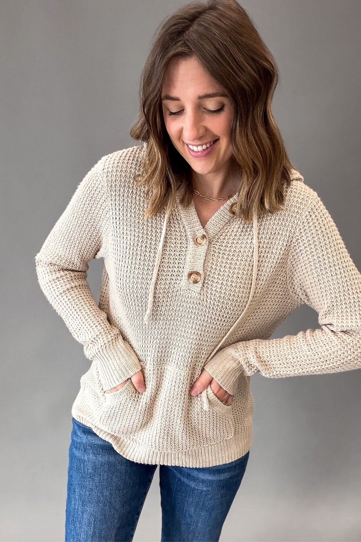 A woman wearing a taupe-colored waffle-knit hoodie sweater with drawstrings and buttons, paired with blue jeans, posing against a solid gray studio background.