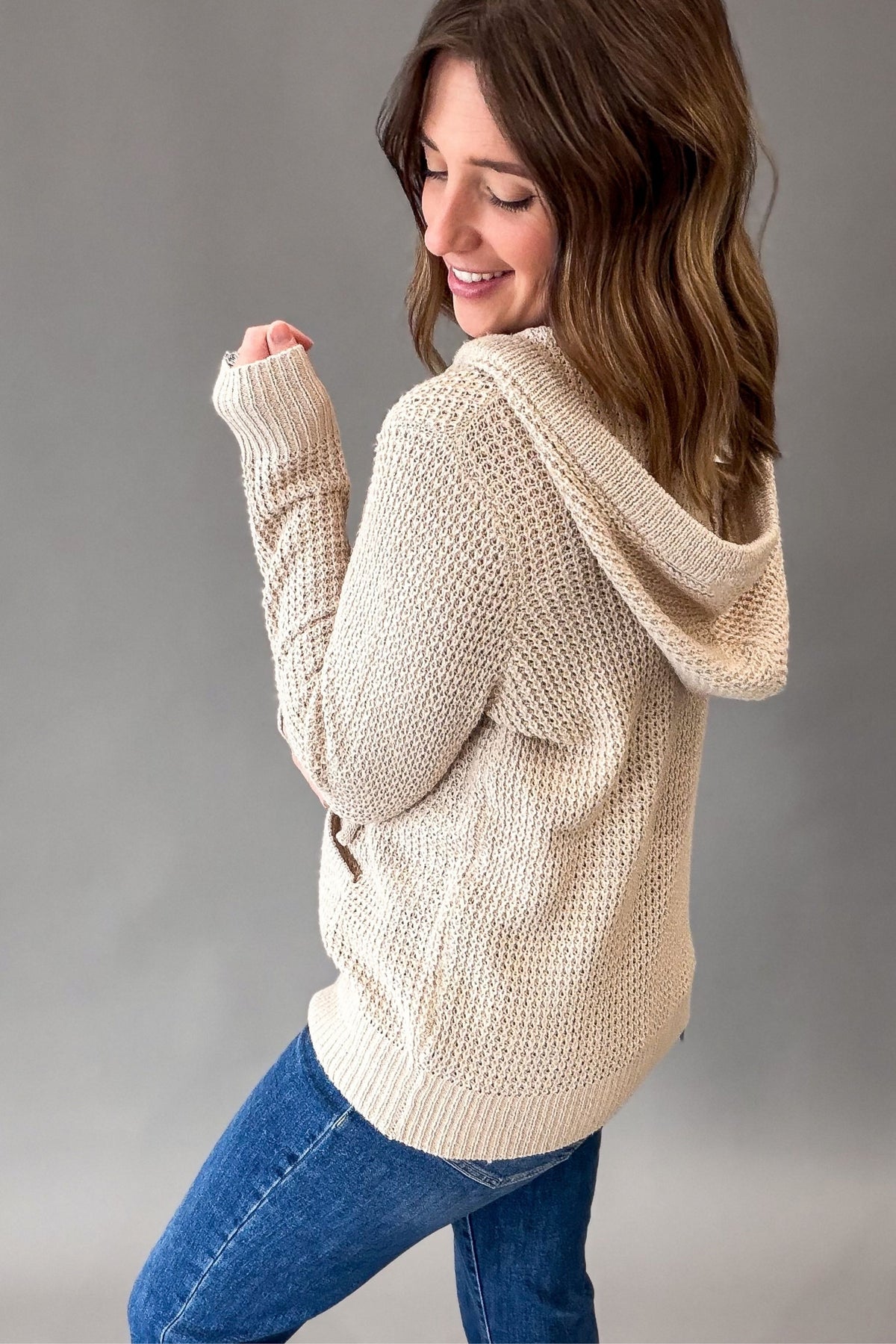 A woman wearing a taupe-colored, chunky knit hooded sweater and blue jeans, photographed from a side-back angle against a neutral gray studio background.