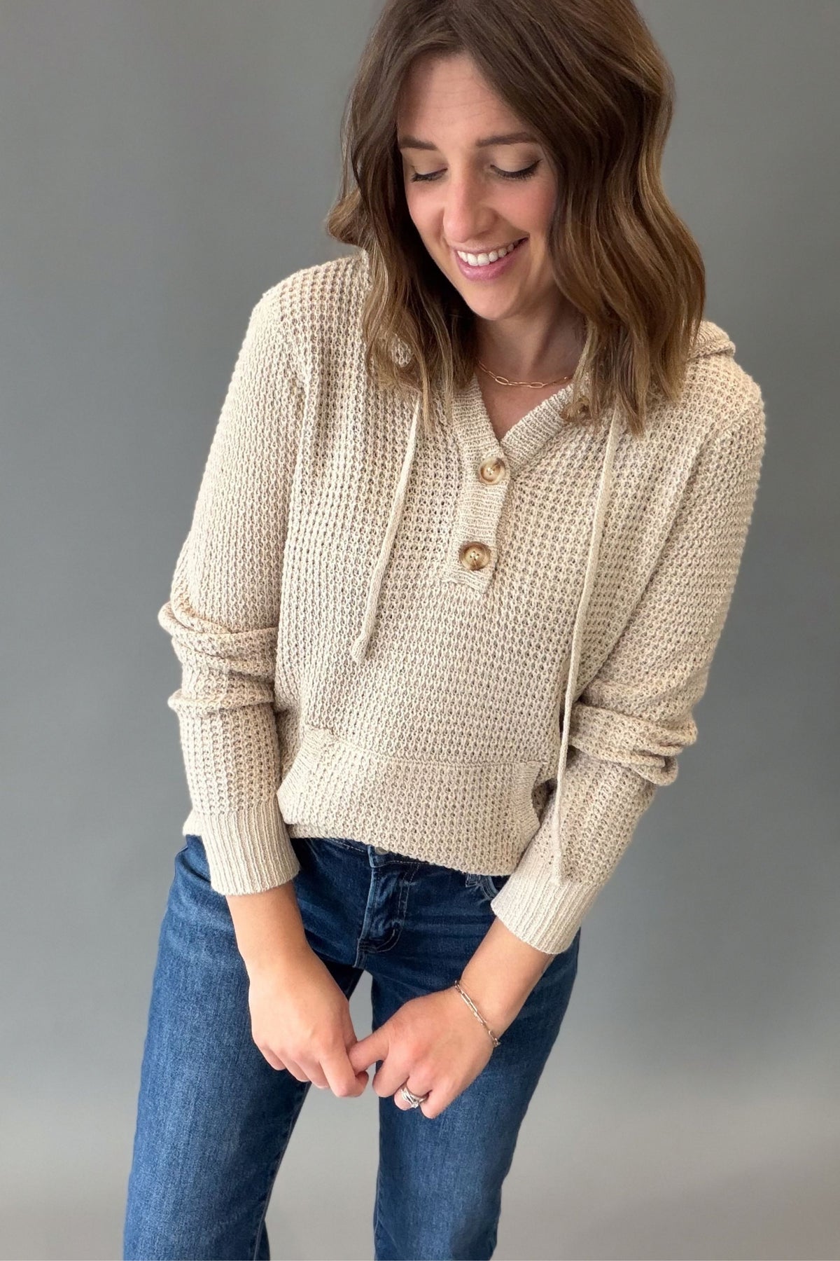 A woman wearing a taupe-colored waffle-knit hoodie with drawstrings and a buttoned placket, paired with blue jeans, standing against a neutral gray studio background.