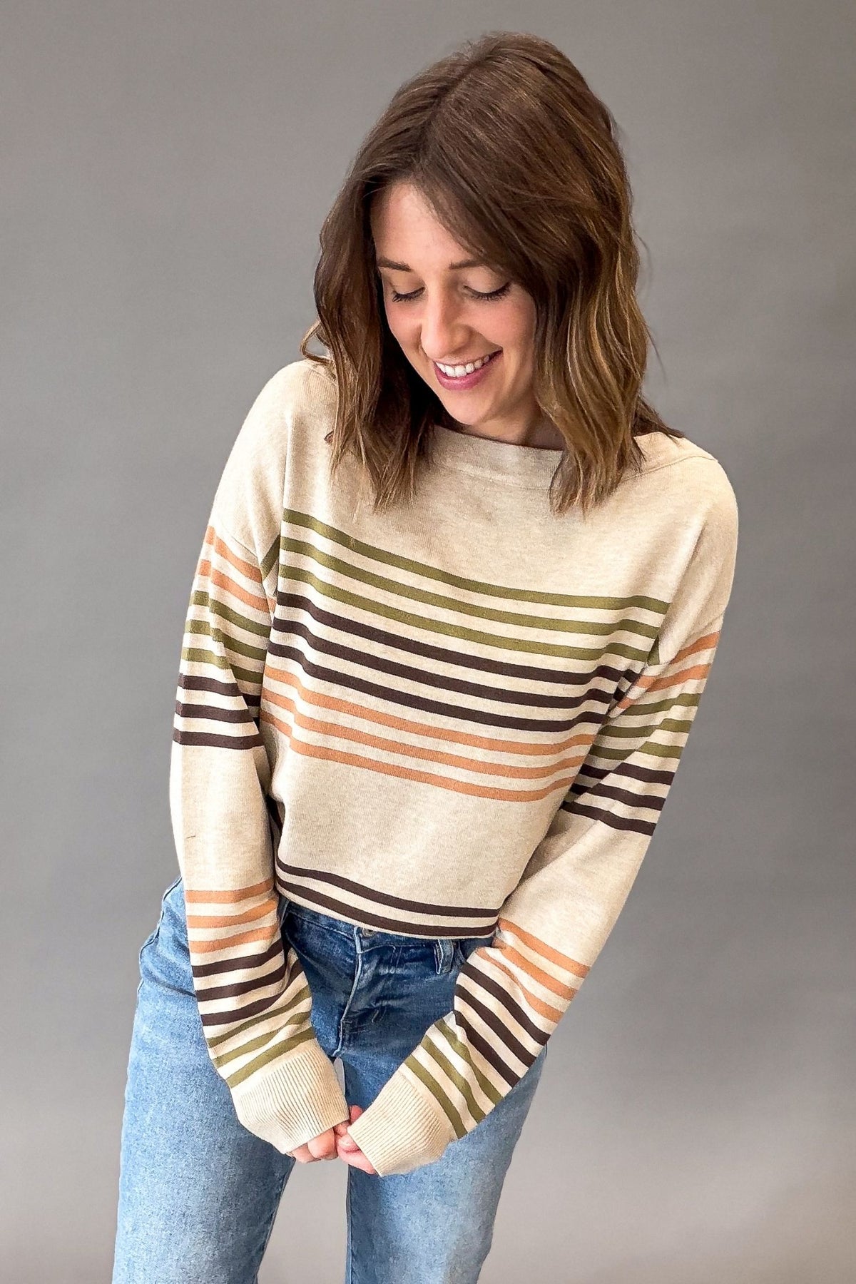 A woman wearing a cream-colored knit sweater with horizontal stripes in olive green, brown, and orange, paired with blue jeans, posing against a solid gray studio background.
