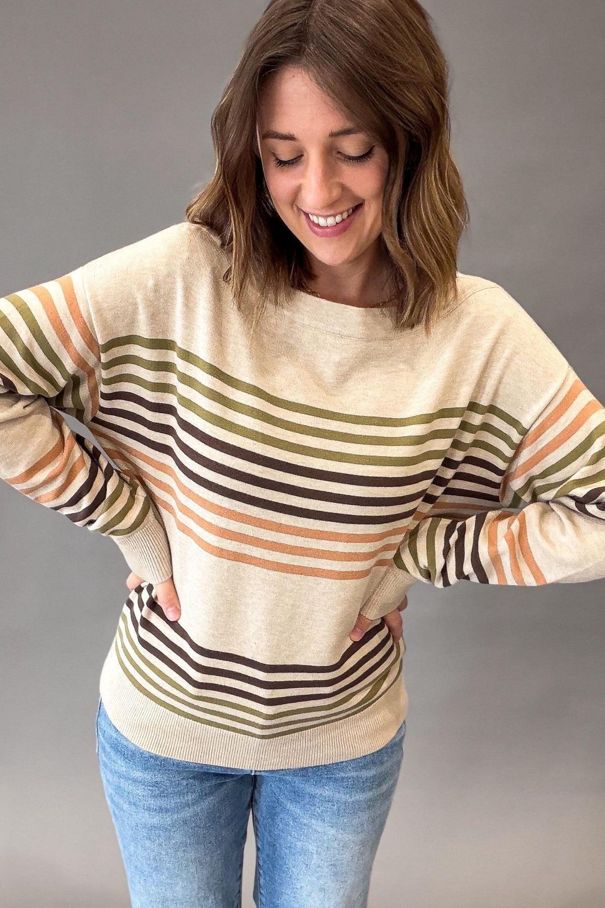 A woman wearing a cream-colored knit sweater with horizontal stripes in olive green, dark brown, and orange, paired with blue jeans, against a solid gray studio background.