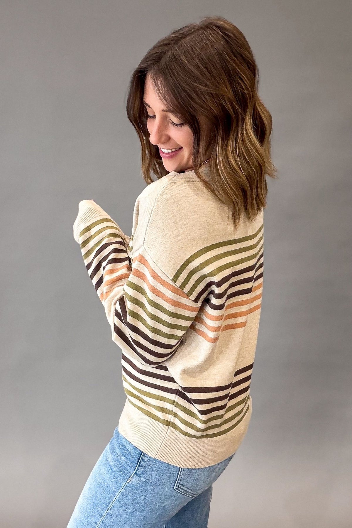 A woman wearing a cream-colored knit sweater with horizontal stripes in olive green, brown, and peach, paired with light blue jeans, photographed against a neutral gray studio background.