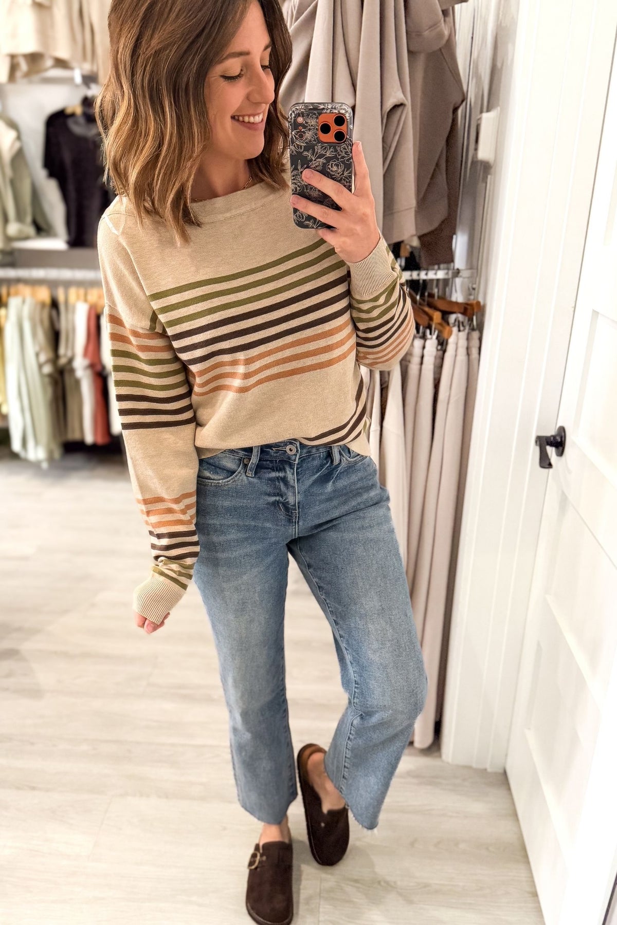 A woman takes a mirror selfie in a clothing store while wearing a beige crewneck sweater with horizontal stripes in olive green, brown, and orange, paired with blue jeans and brown loafers.