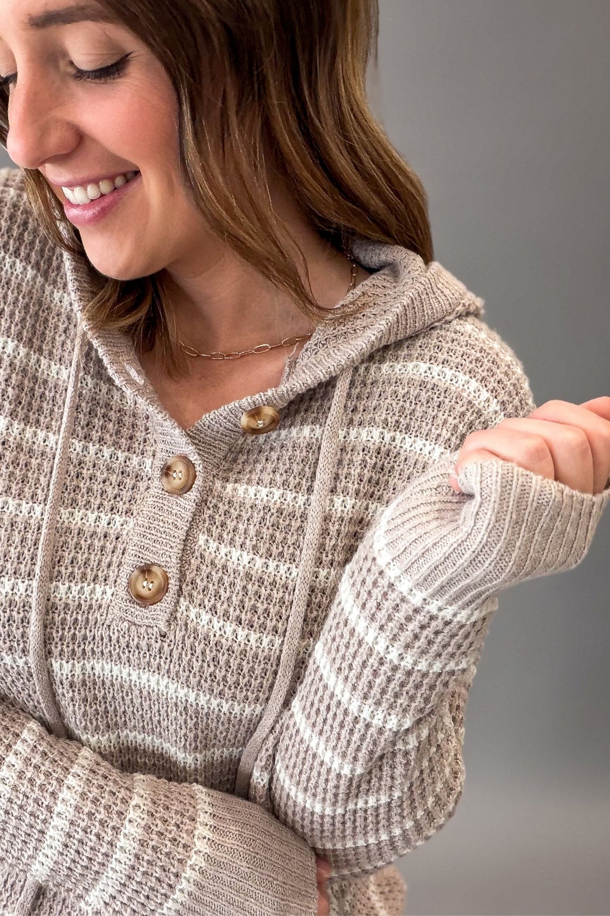 A close-up of a woman wearing a taupe and off-white waffle-knit hooded sweater with brown buttons, shown against a plain gray background.