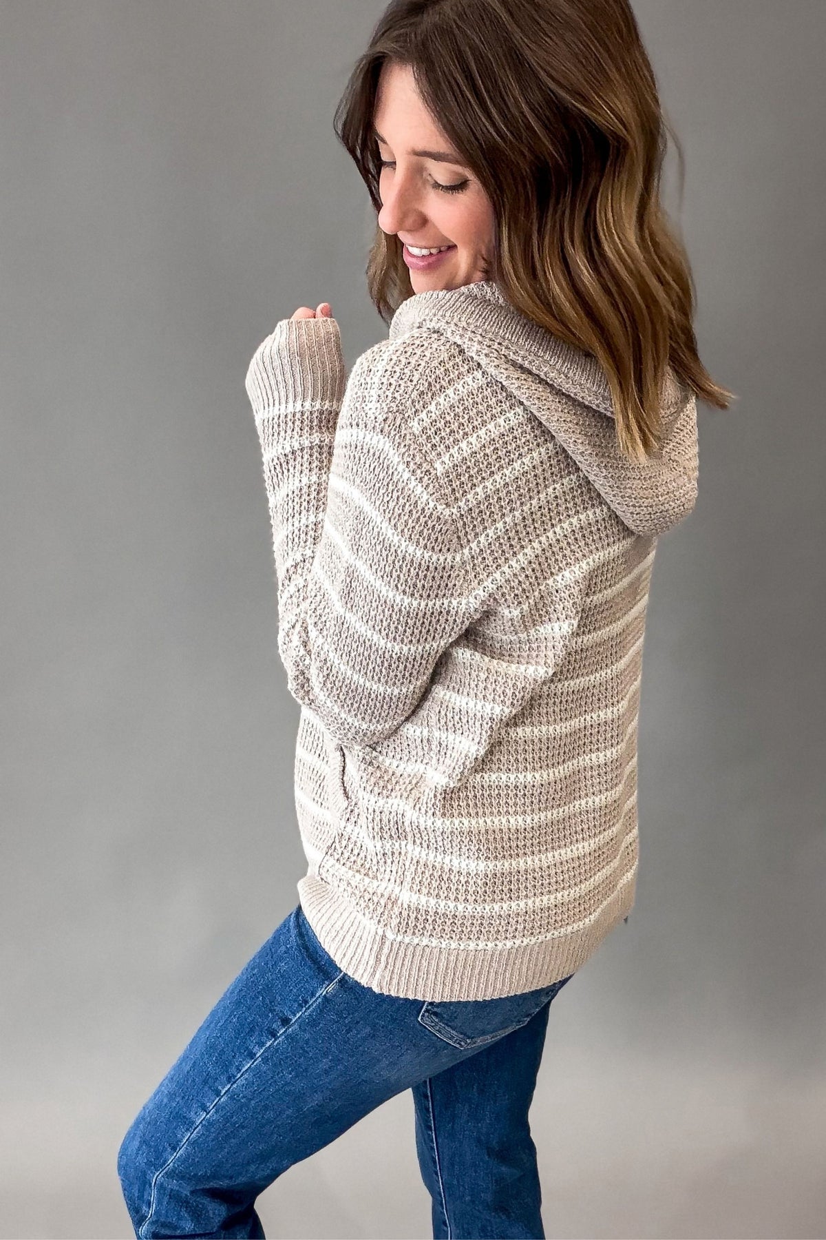 A woman wearing a chunky knit hooded sweater with horizontal taupe and cream stripes, paired with blue jeans, photographed against a neutral gray studio background.
