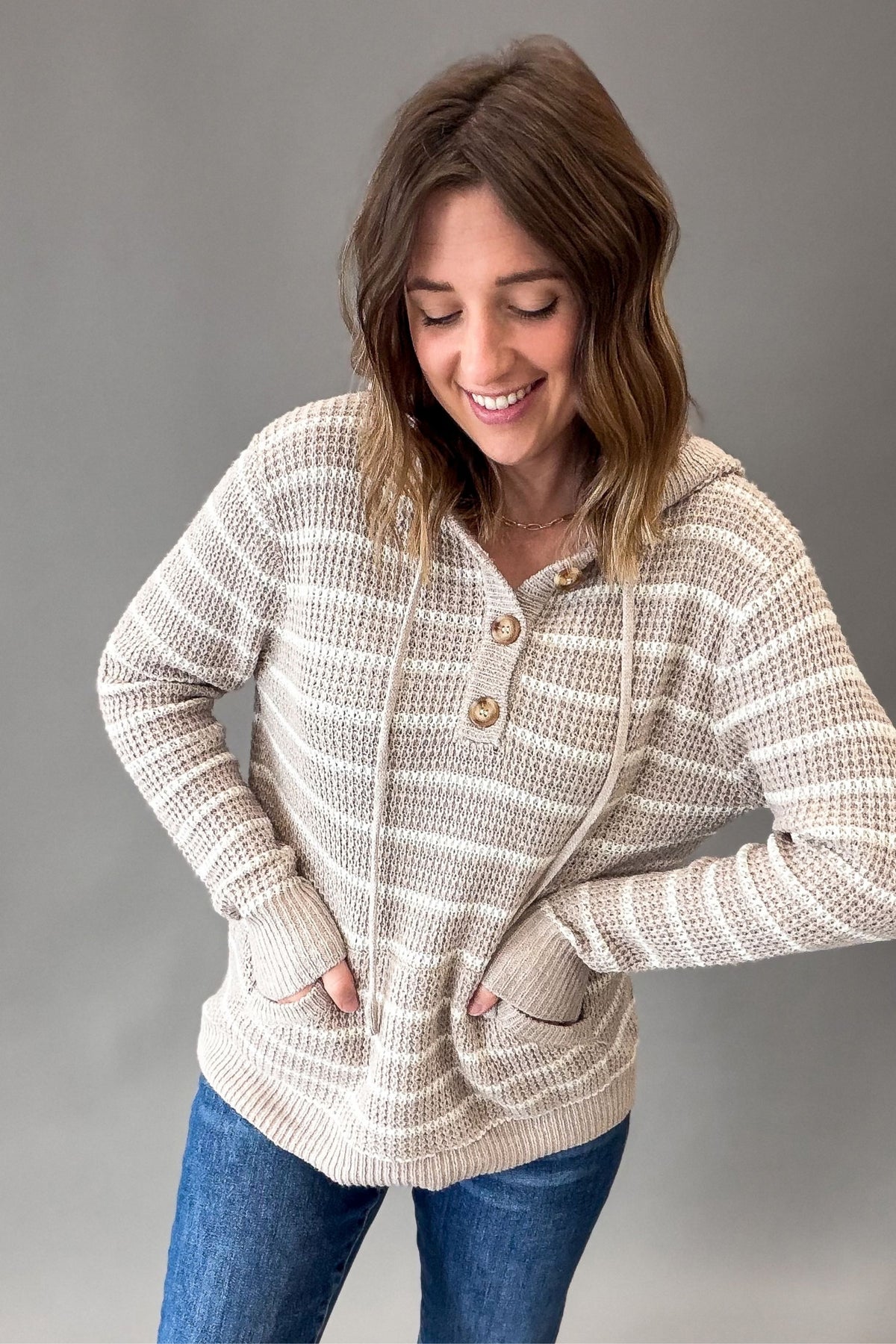 A woman wearing a taupe and cream striped knit cardigan with a buttoned henley and front pockets, paired with blue jeans, against a solid gray studio background.