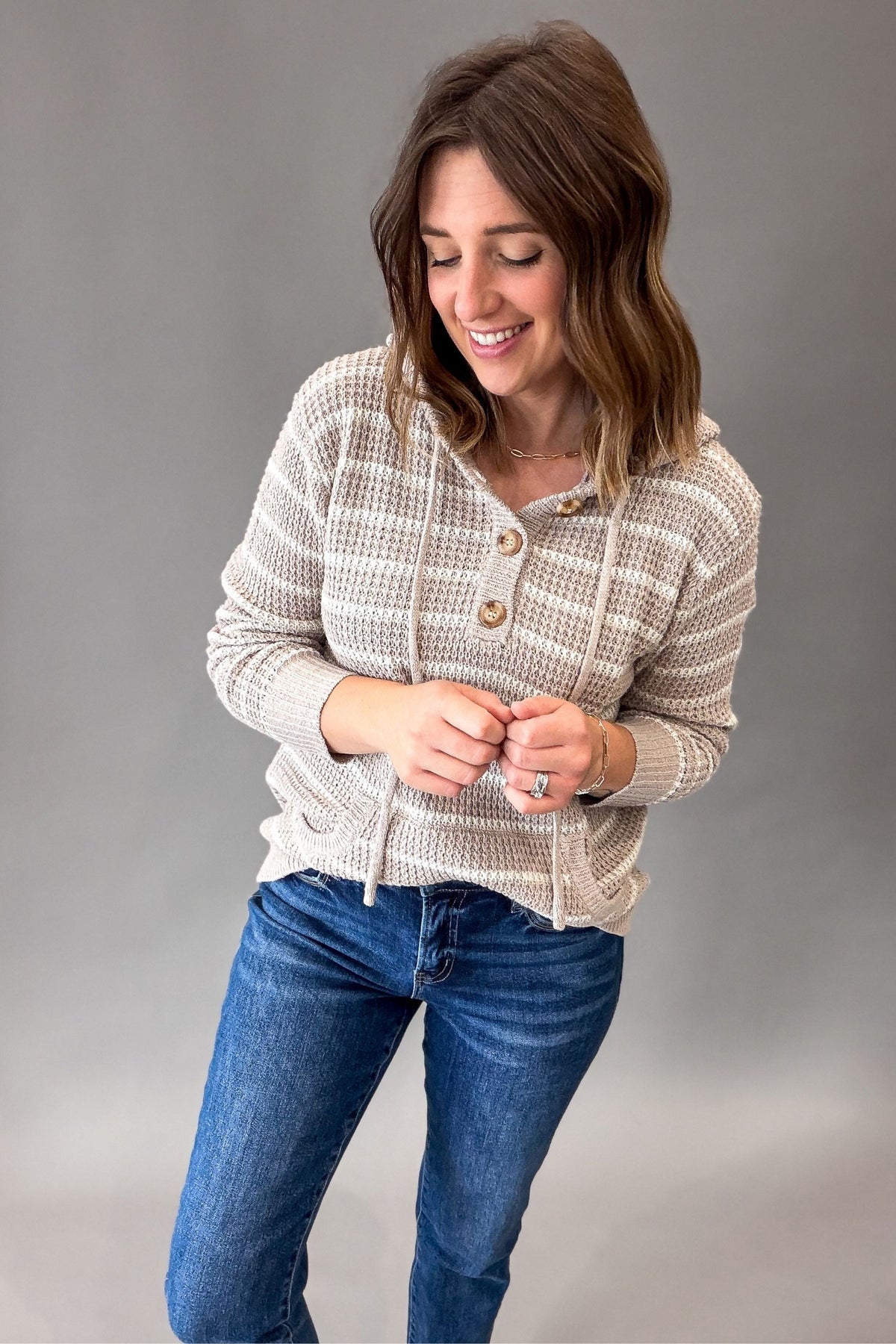A woman wearing a taupe and white textured knit hooded sweater with drawstrings and buttons, paired with blue jeans, standing against a neutral gray studio background.