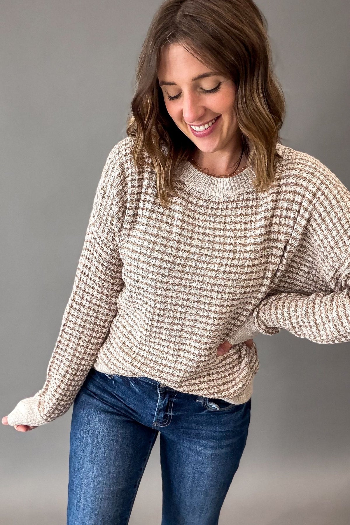 A woman wearing a beige and cream textured knit sweater with blue jeans, posing against a solid gray studio background.